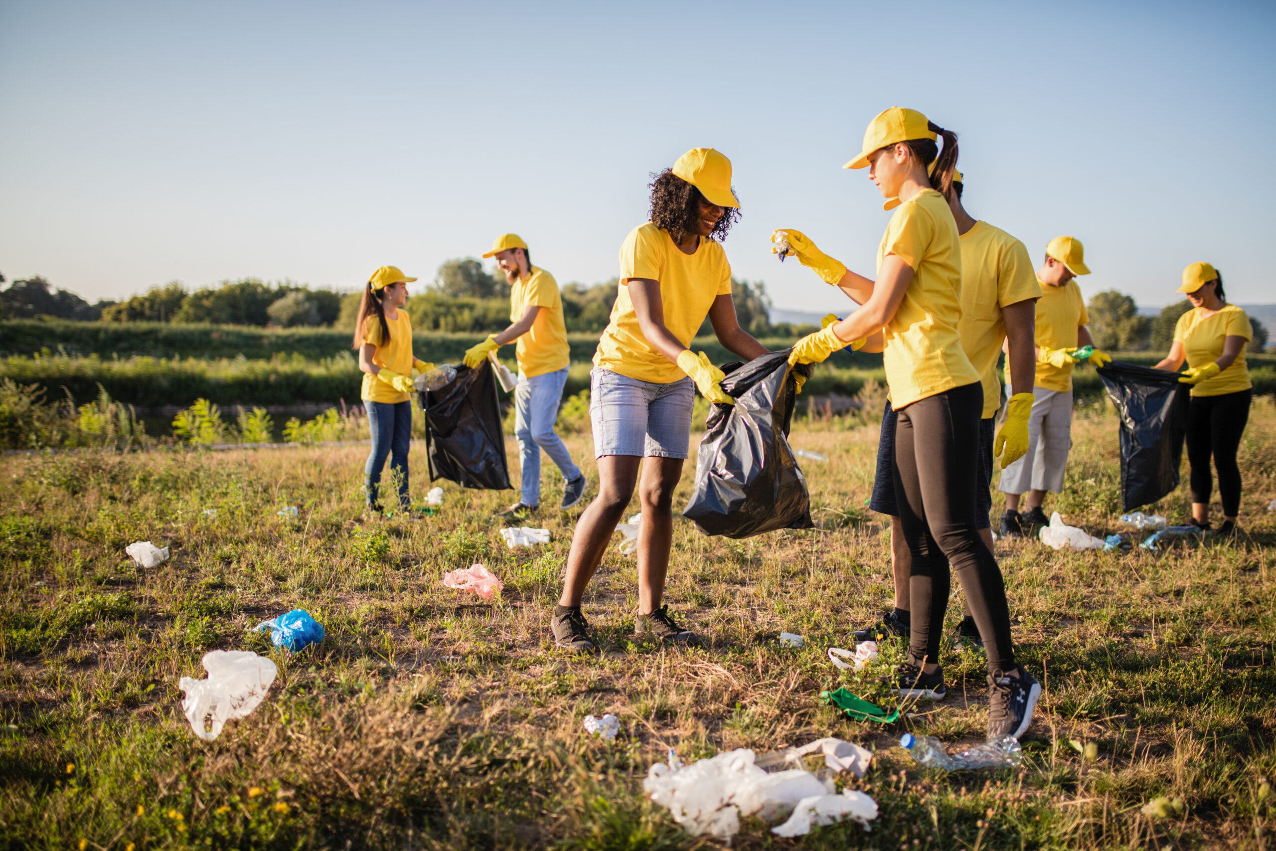 A large group of people wearing yelow shirts and hats, picking up trash in a big field.