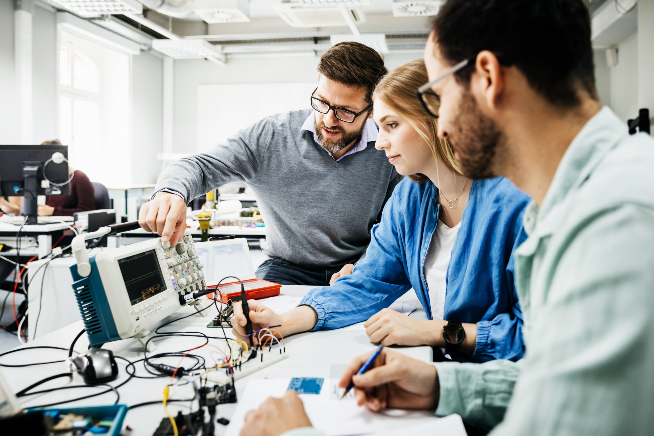 A professor shows two students how to work with electronic equipment in a classroom.