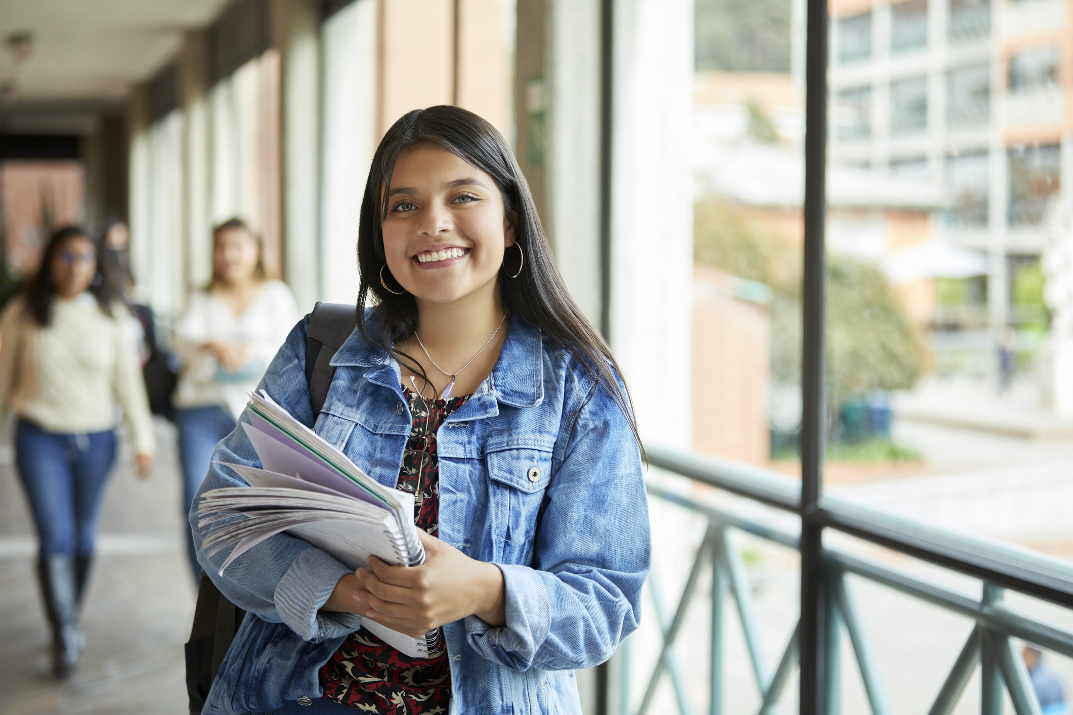 Happy college transfer student holding notebooks and wearing a backpack stops to smile at the camera while walking to class on campus.