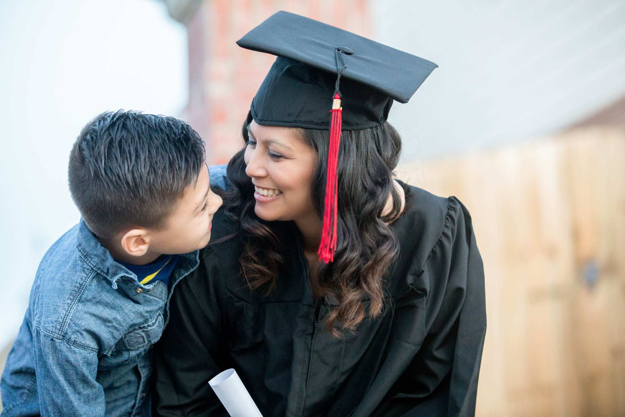 A woman dressed in college graduation regalia leans over and smiles at her young son.