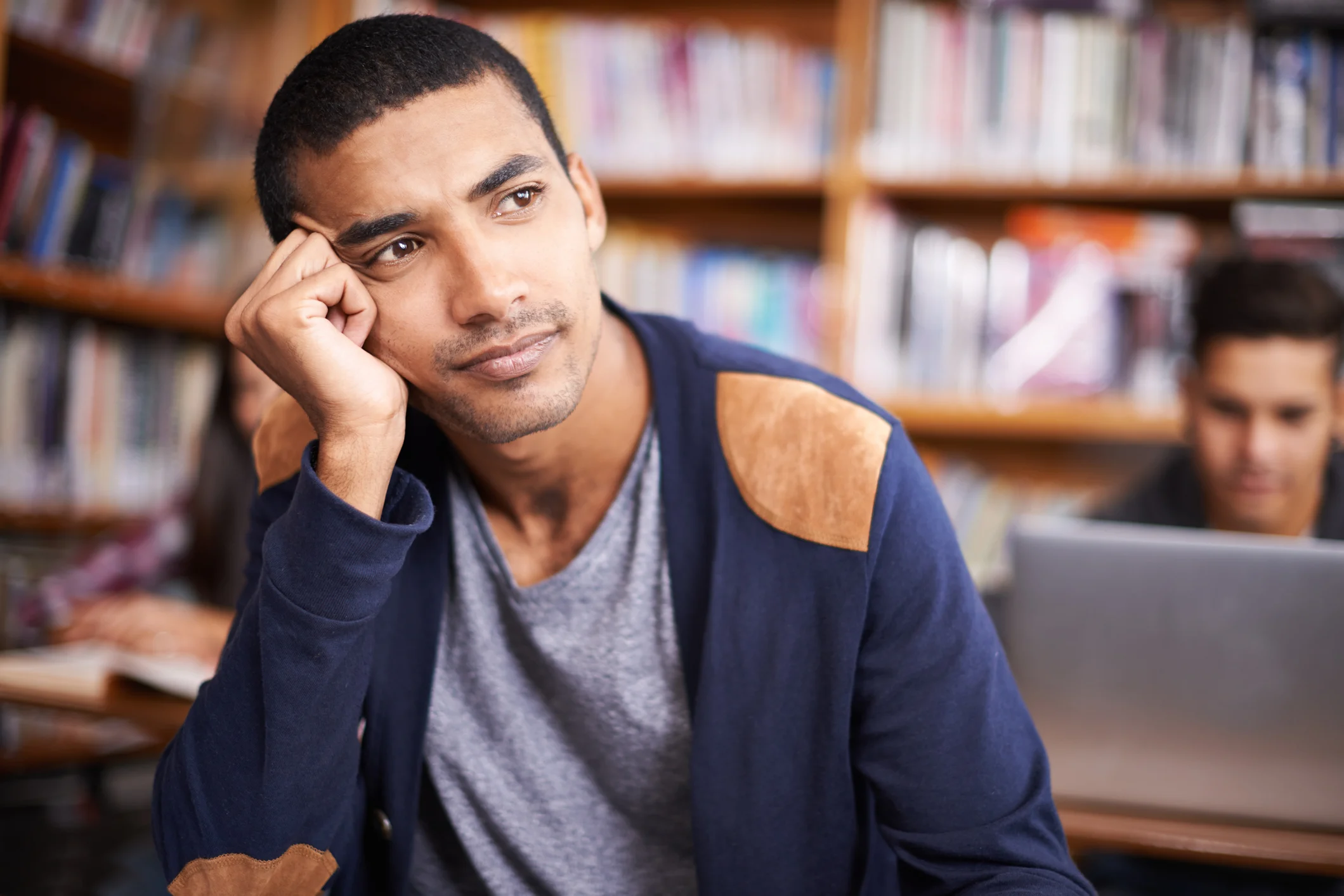 A young man thinks in a library, his head resting in his palm.