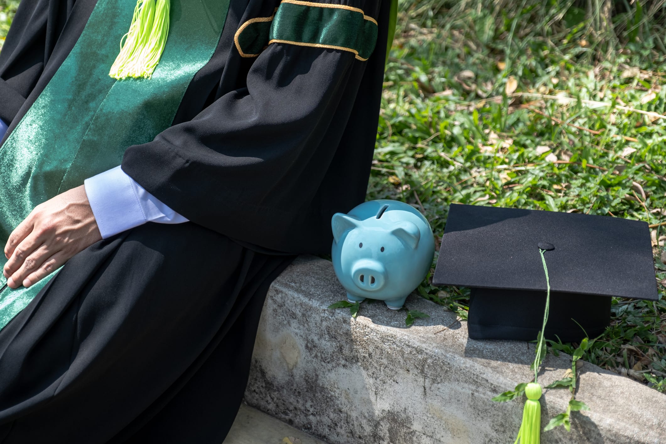 A college graduate sits outside beside a blue piggy bank and a graduation cap.