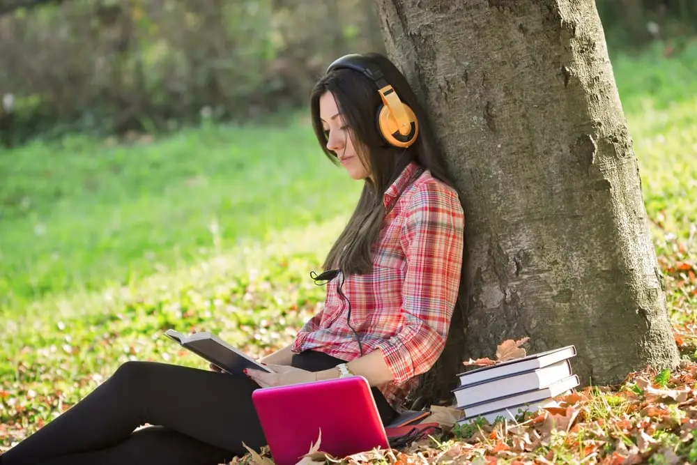 A young woman wearing headphones sits with her back against a tree as she reads a book for school.