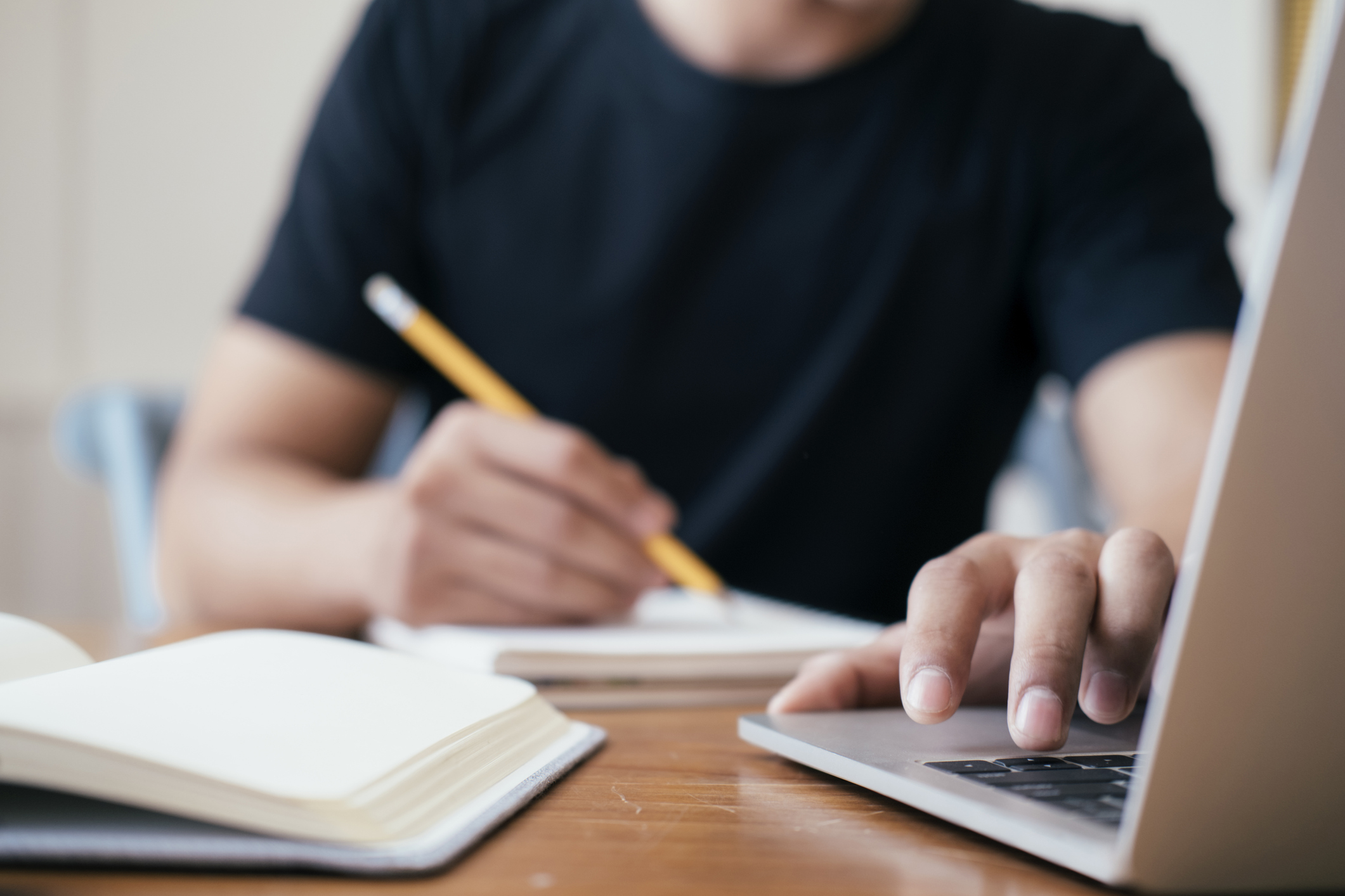 High school student taking notes on a notepad while using his laptop computer to search for colleges.