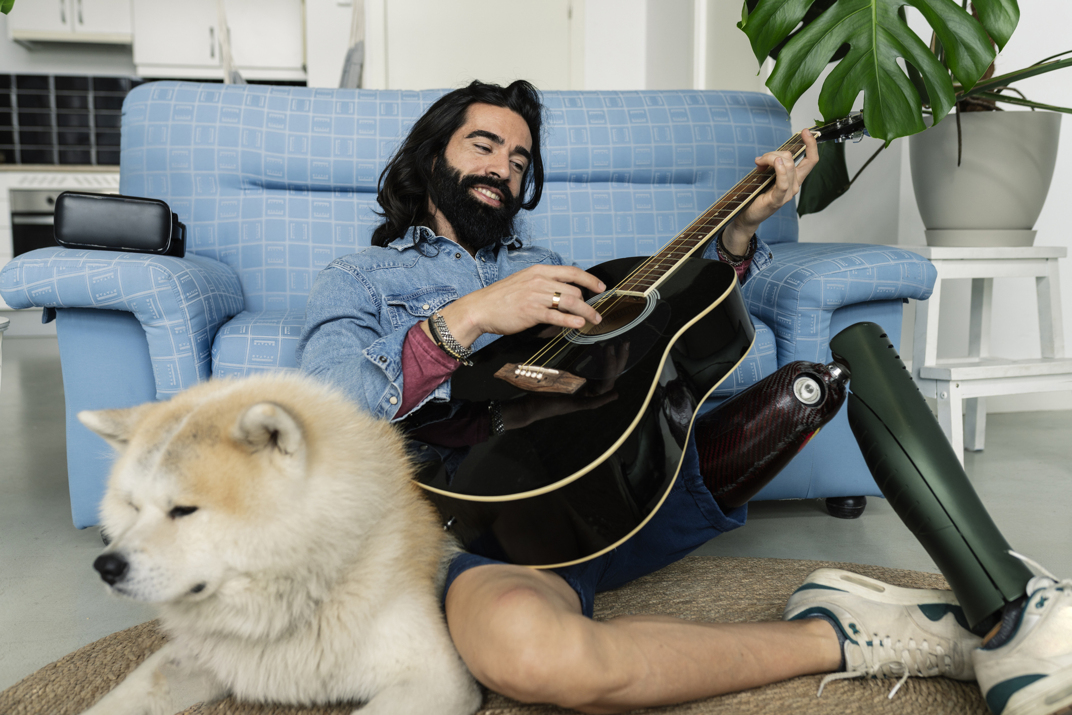 Smiling college student with a prosthetic leg plays a guitar while sitting next to his dog in his apartment living room.