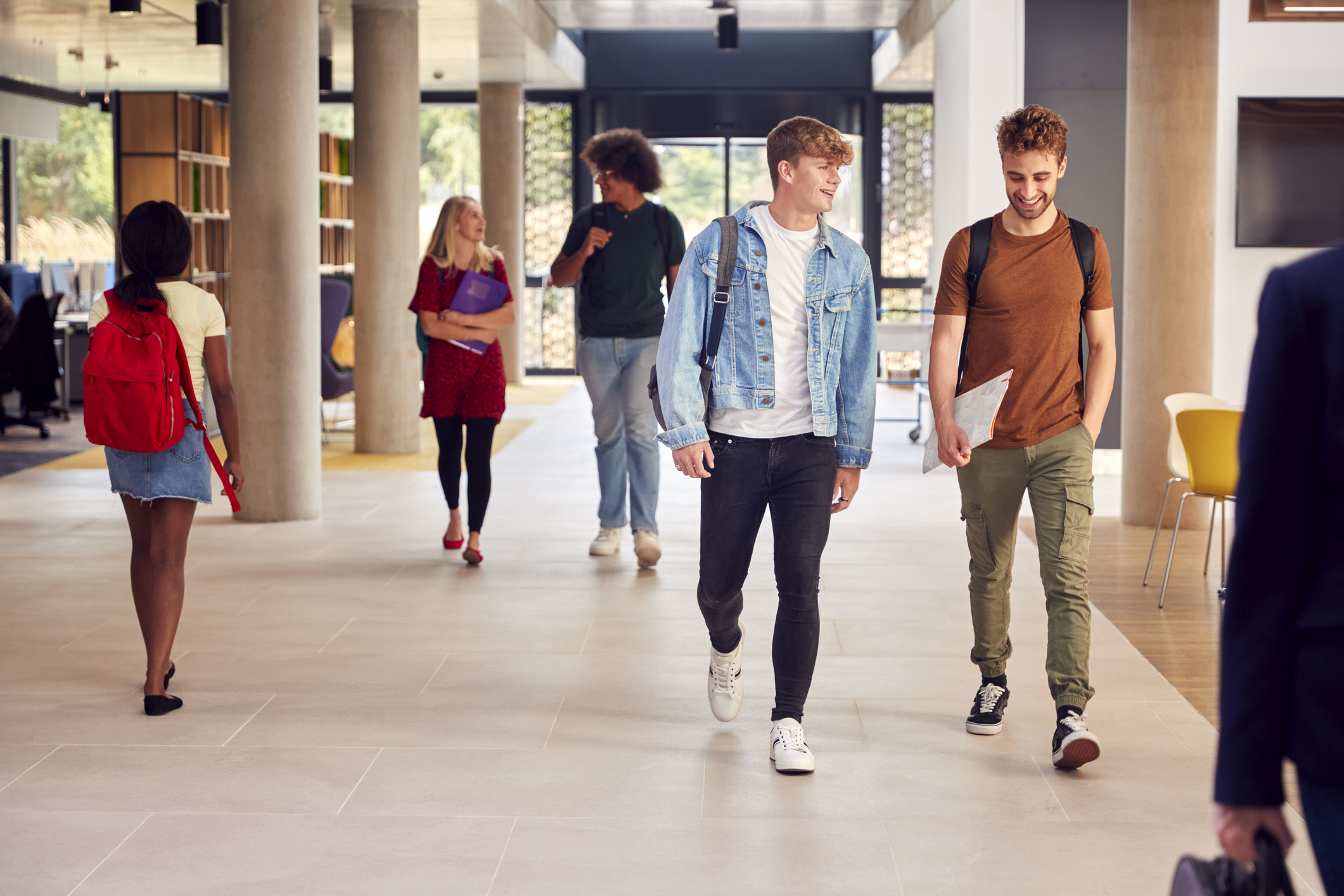 Two Male Students In Busy University Or College Building Talking As They Walk Along Corridor