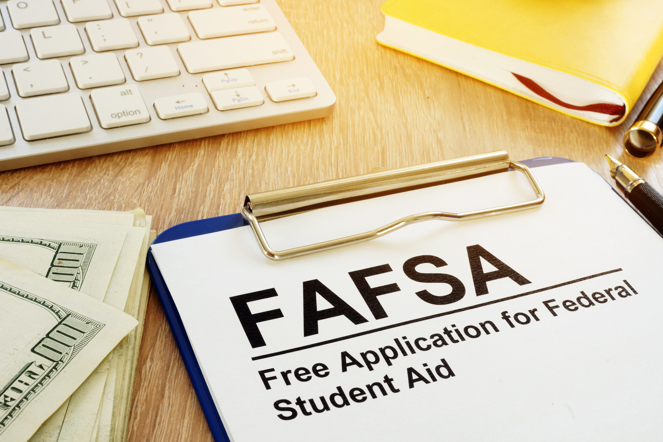 A clipboard with a piece of paper in it reading "FAFSA: Free Application for Federal Student Aid" lies on a desk next to a keyboard and a small pile of cash.