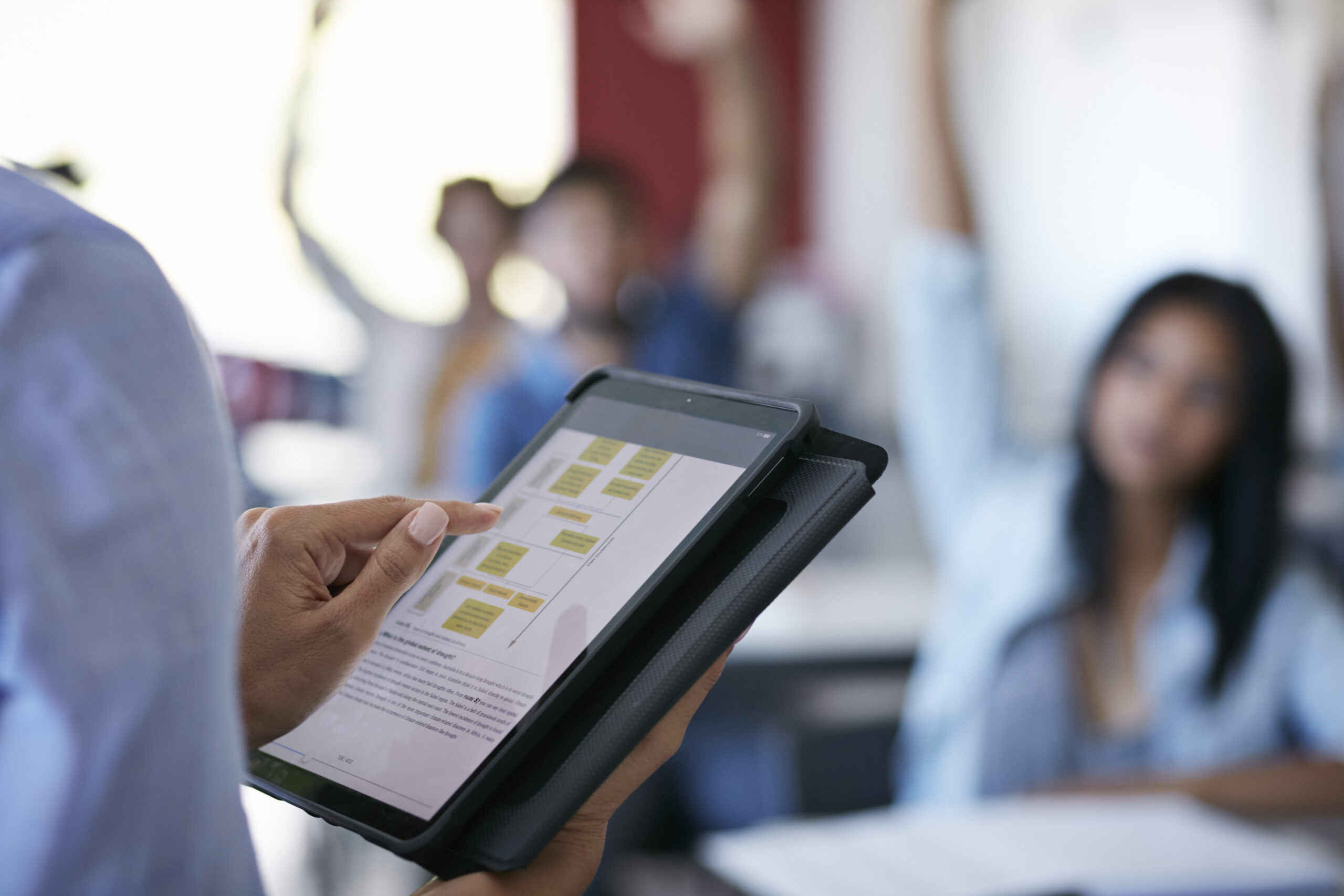 Close-up of teacher using tablet in class, students with raised hands in back