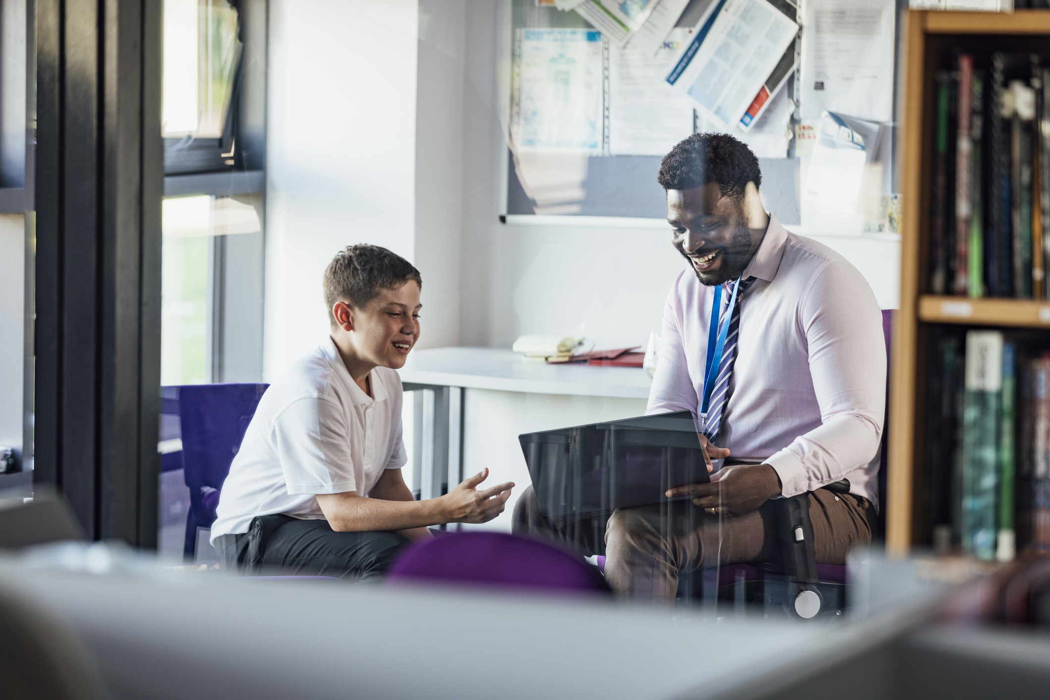 A male secondary school student sitting in an office in the school, having a meeting with his teacher. The teacher is showing him something on his laptop while they both smile.