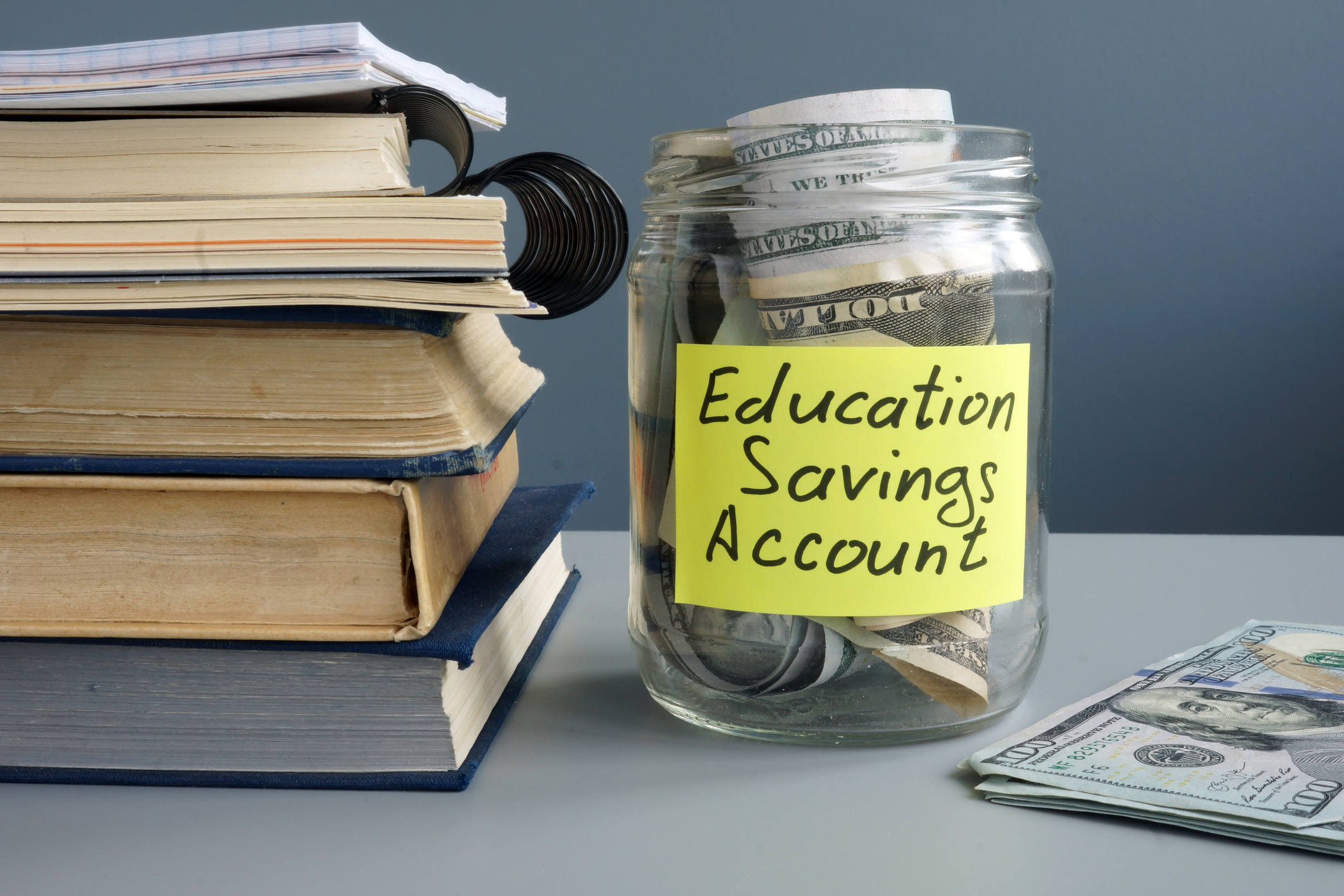 A stack of books sits beside a jar of cash with a yellow post-it label reading "Education Savings Account."