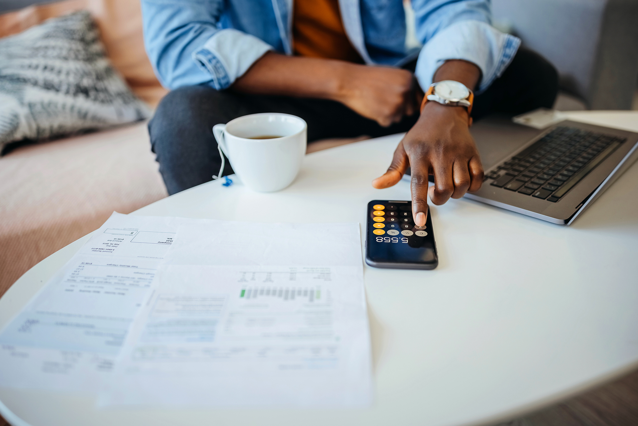 A college student sits in his living room with paper bills and his laptop open on his desk. He is using the calculator app on his mobile phone to go over his finances.