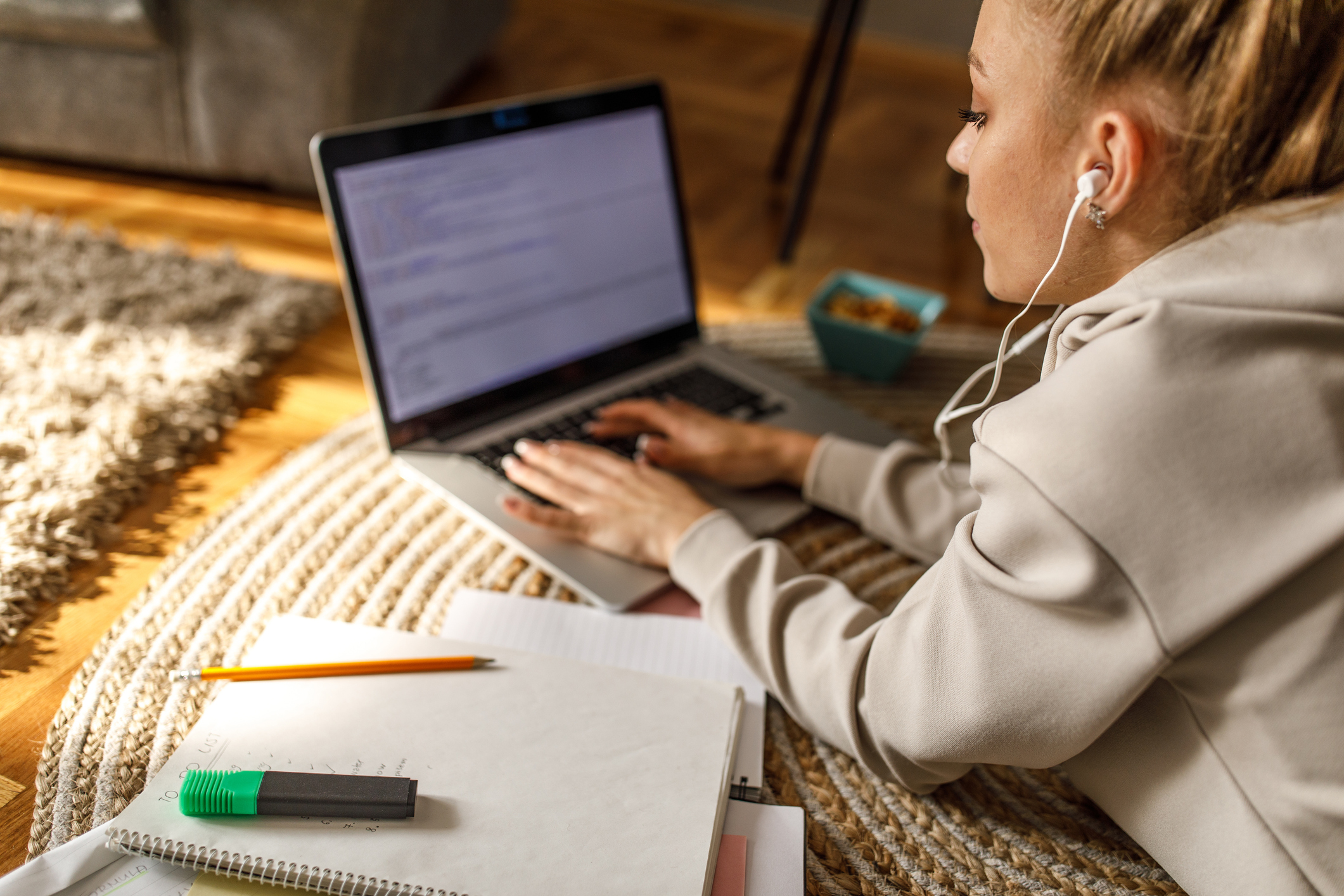 High school student lying on the rug in her living room types her college application essay on her laptop.