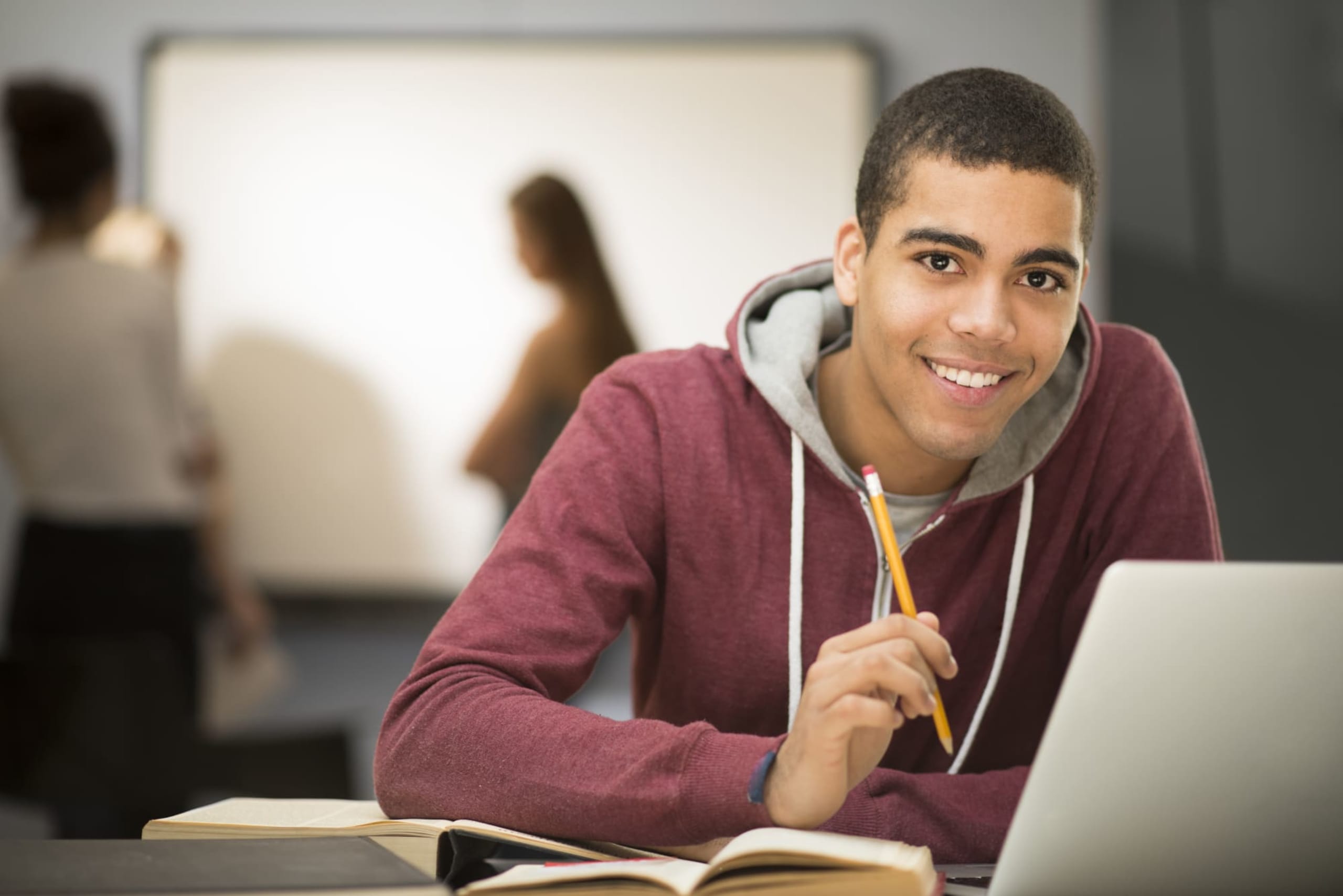 Photo of happy student sitting in front of their laptop in a class with a white board and 2 classmates behind them.