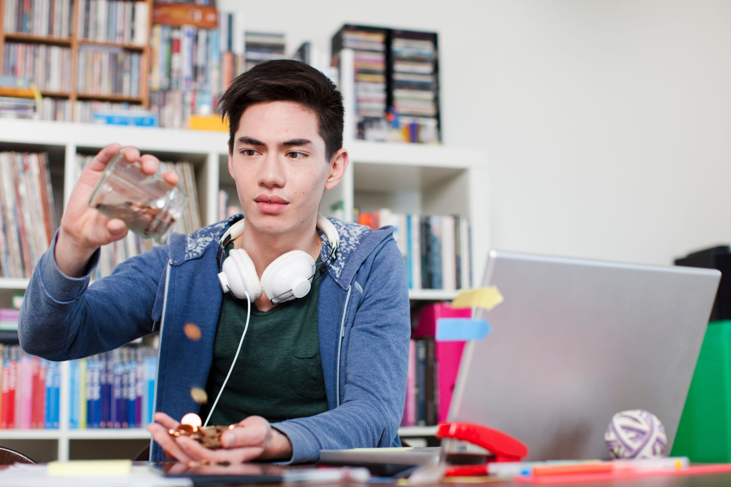 Young man emptying jar of coins into their hand while sitting at a desk with a laptop open