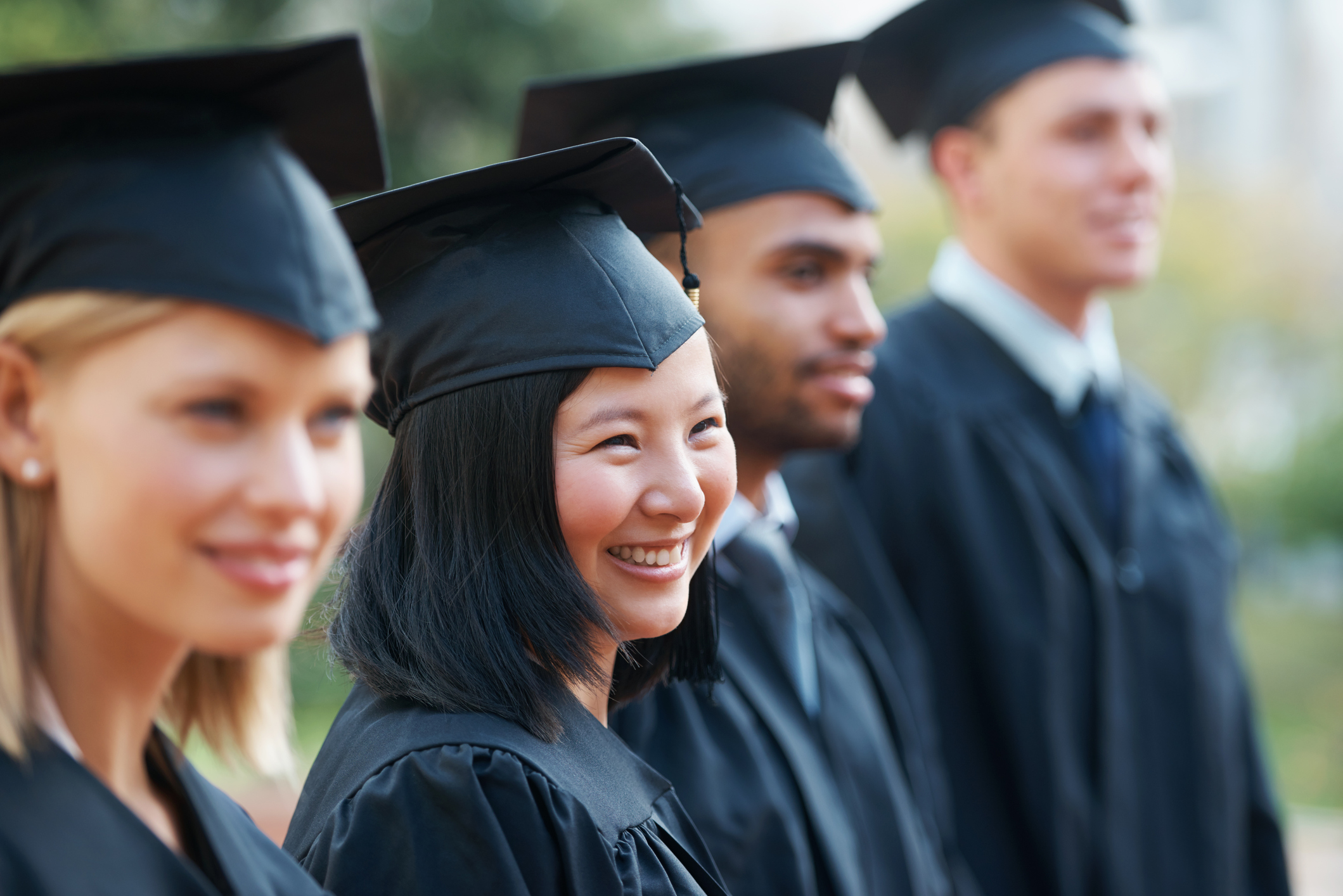 Young college students wearing graduation caps and gowns hold their diplomas while standing in a row and smiling at an outdoor graduation ceremony.