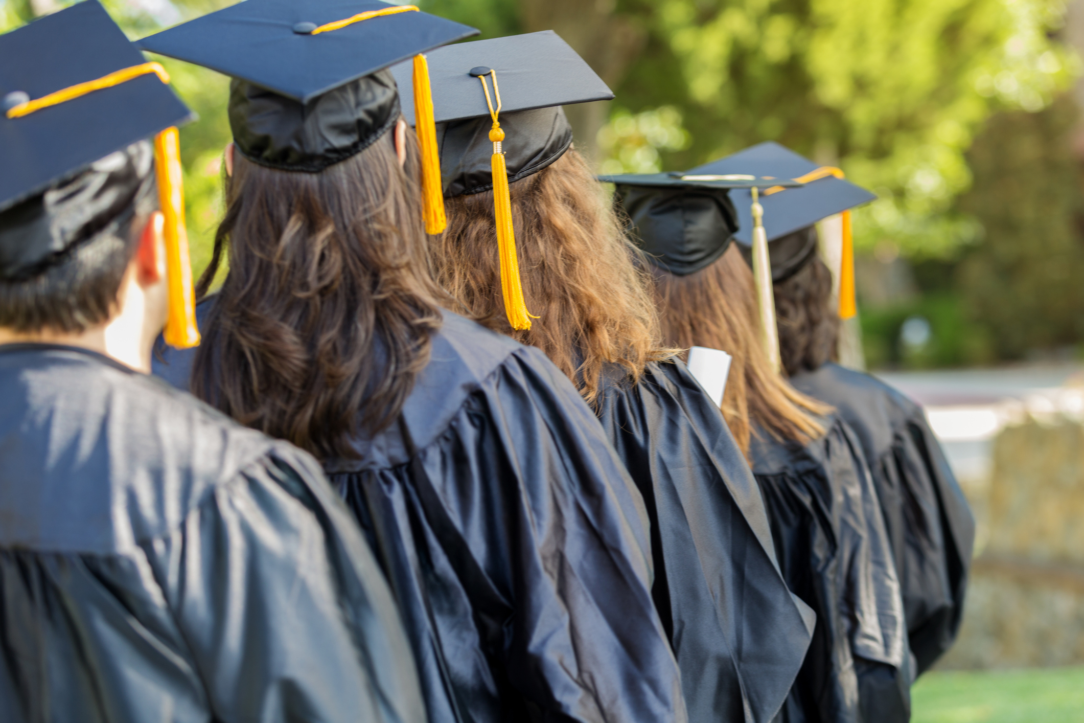 A group of college graduates wearing black caps and gowns stand in line waiting for their diplomas during an outdoor graduation ceremony.