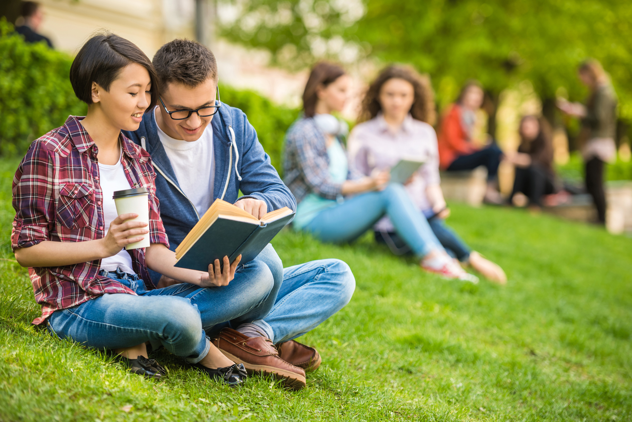 Happy college couple sitting next to each other on a campus lawn, reading from a book while drinking coffee together.