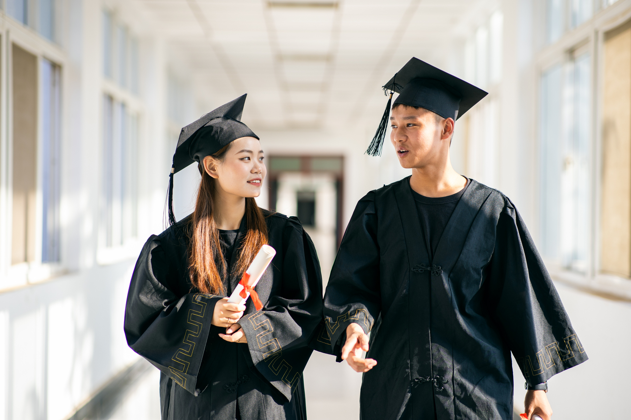 Two Chinese college students dressed in graduation gowns and caps walk through a university hallway building with diplomas in hand.