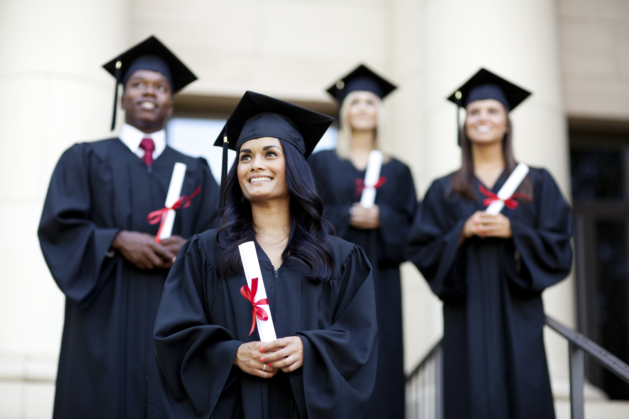 A multicultural group of four college students dressed in graduation gowns and caps stands outside on the steps of a university building with diplomas in their hands.