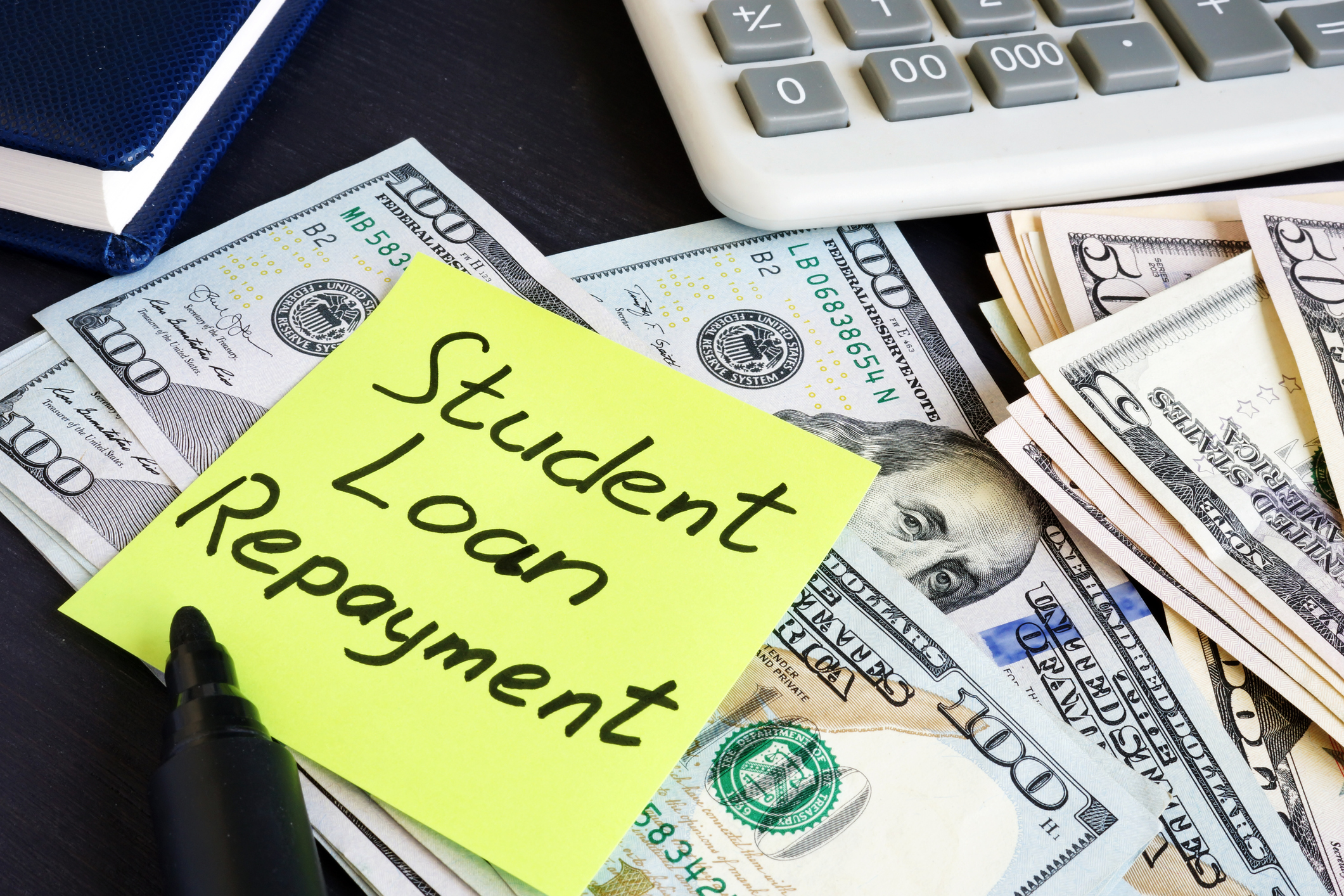 A post-it note with "student loan repayment" written on it laying on top of a stack of U.S. $100 bills.