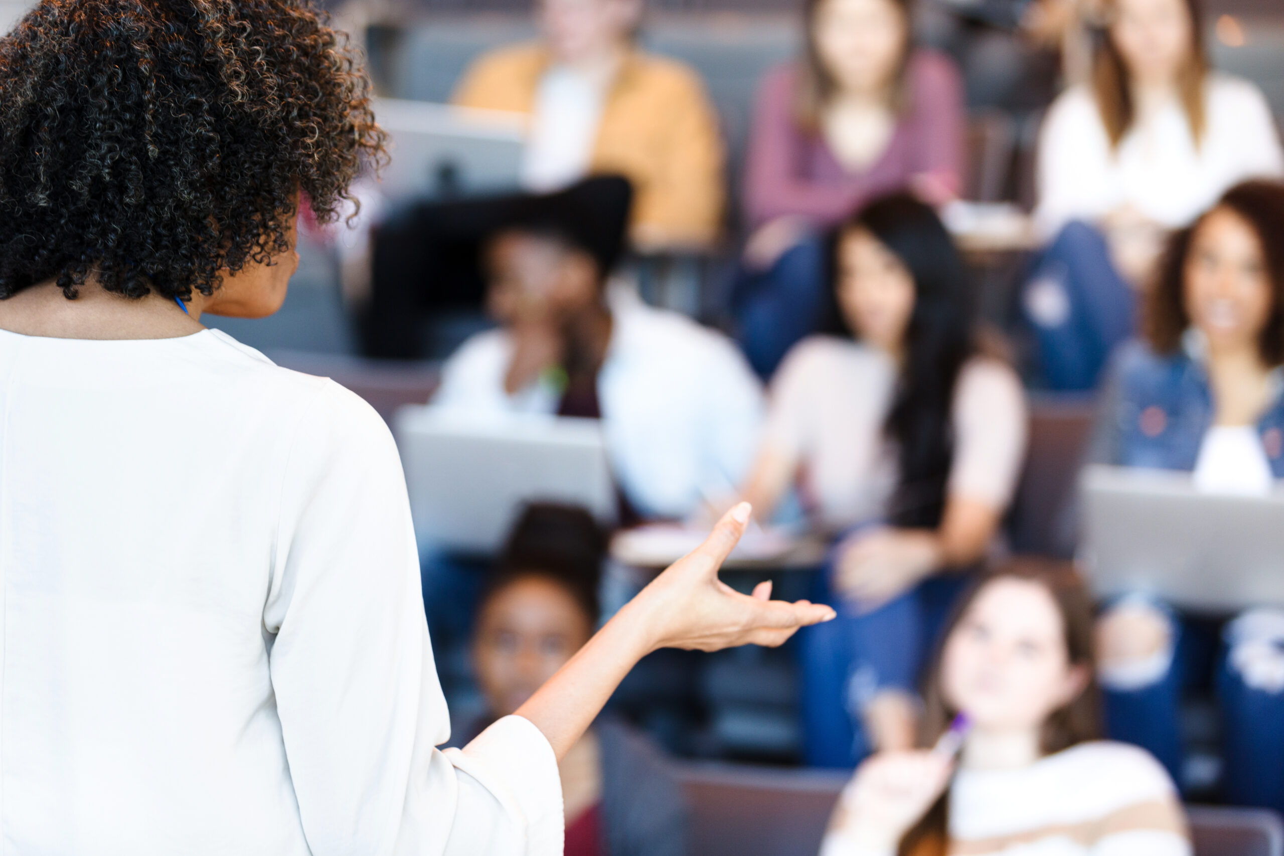 An unrecognizable mature adult female professor gestures while teaching a diverse group of college students.