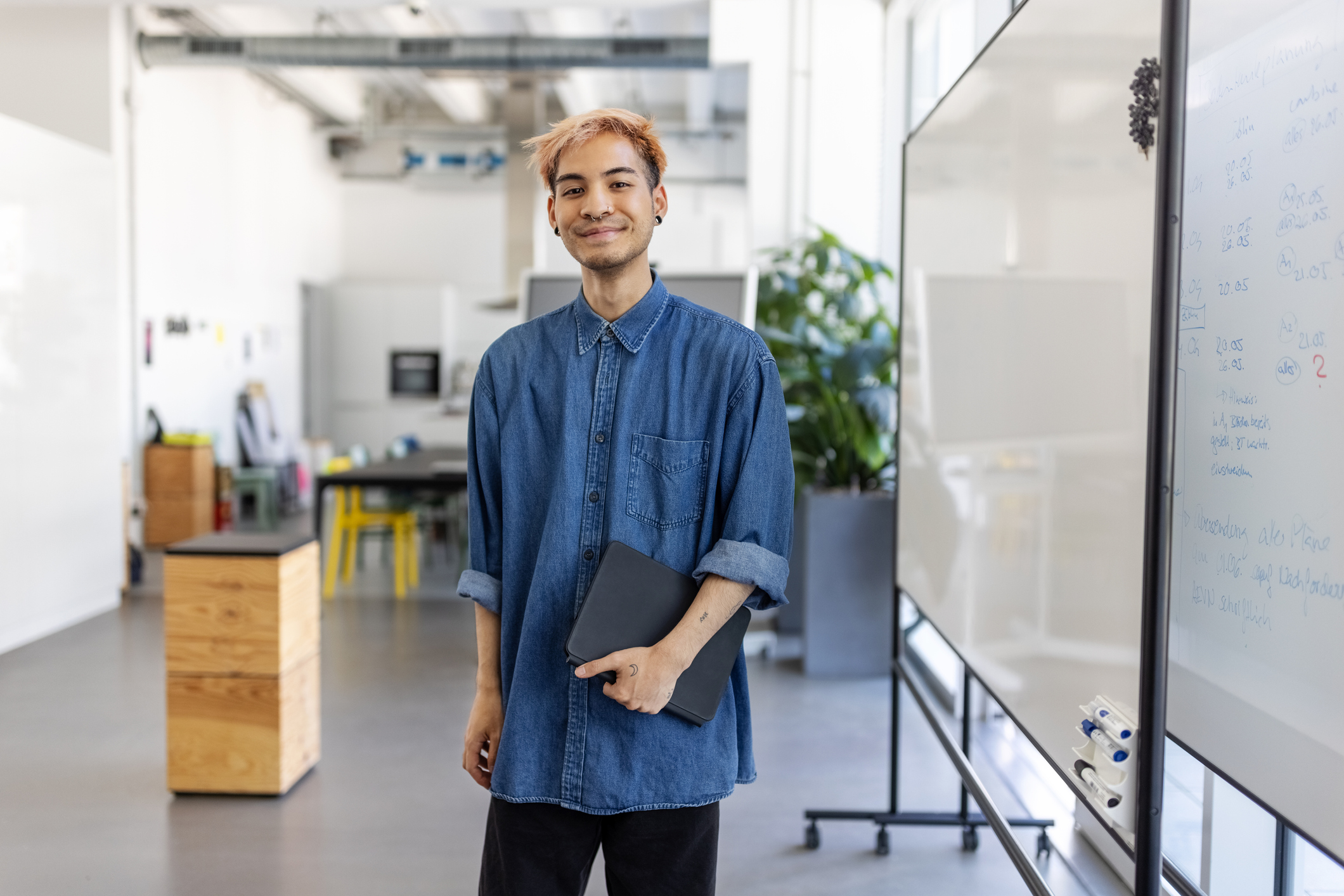 A young professional stands in the center of an office smiling and holding a notebook.