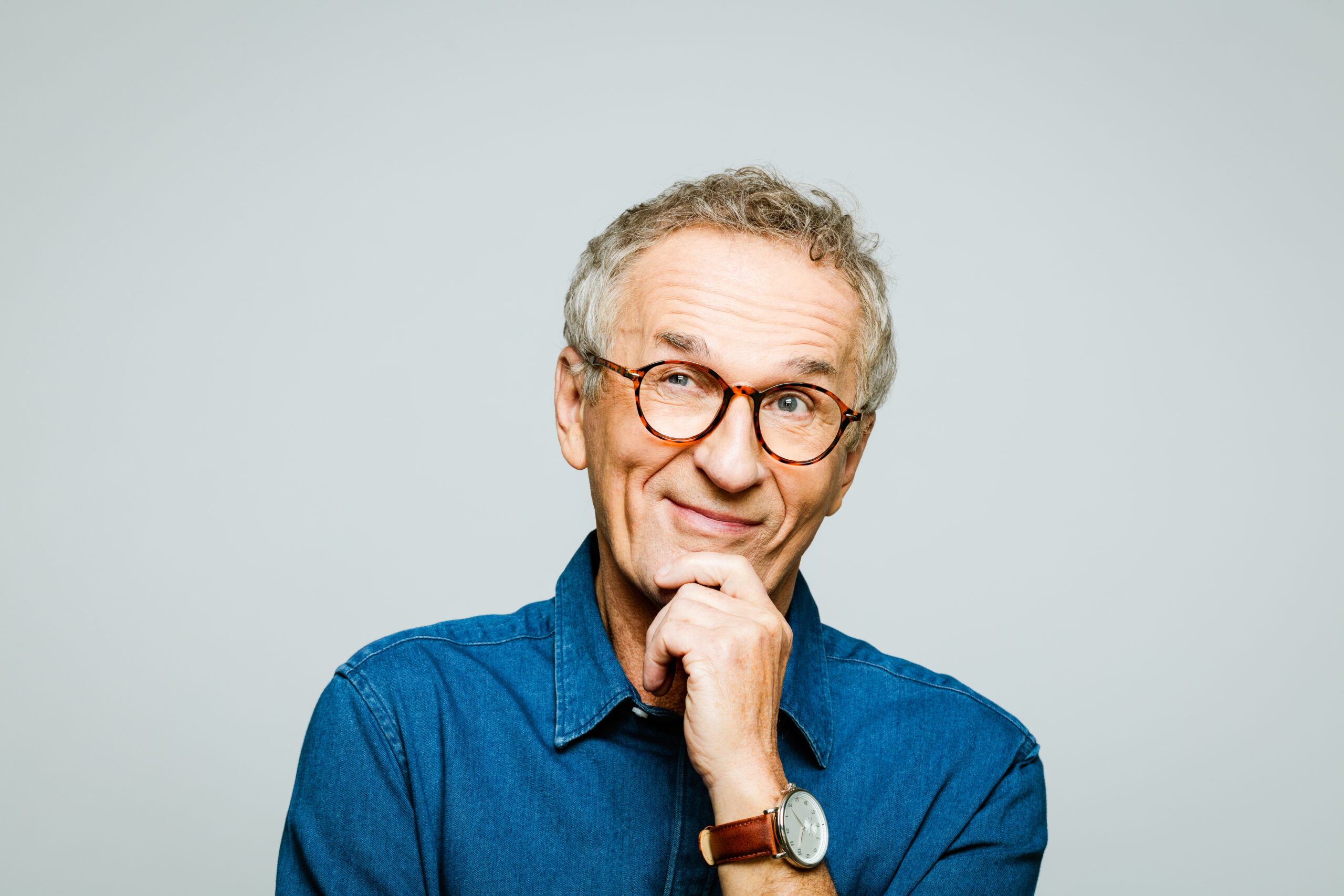 Portrait of elderly man wearing white denim shirt and glasses looking away and smiling with hand on chin.