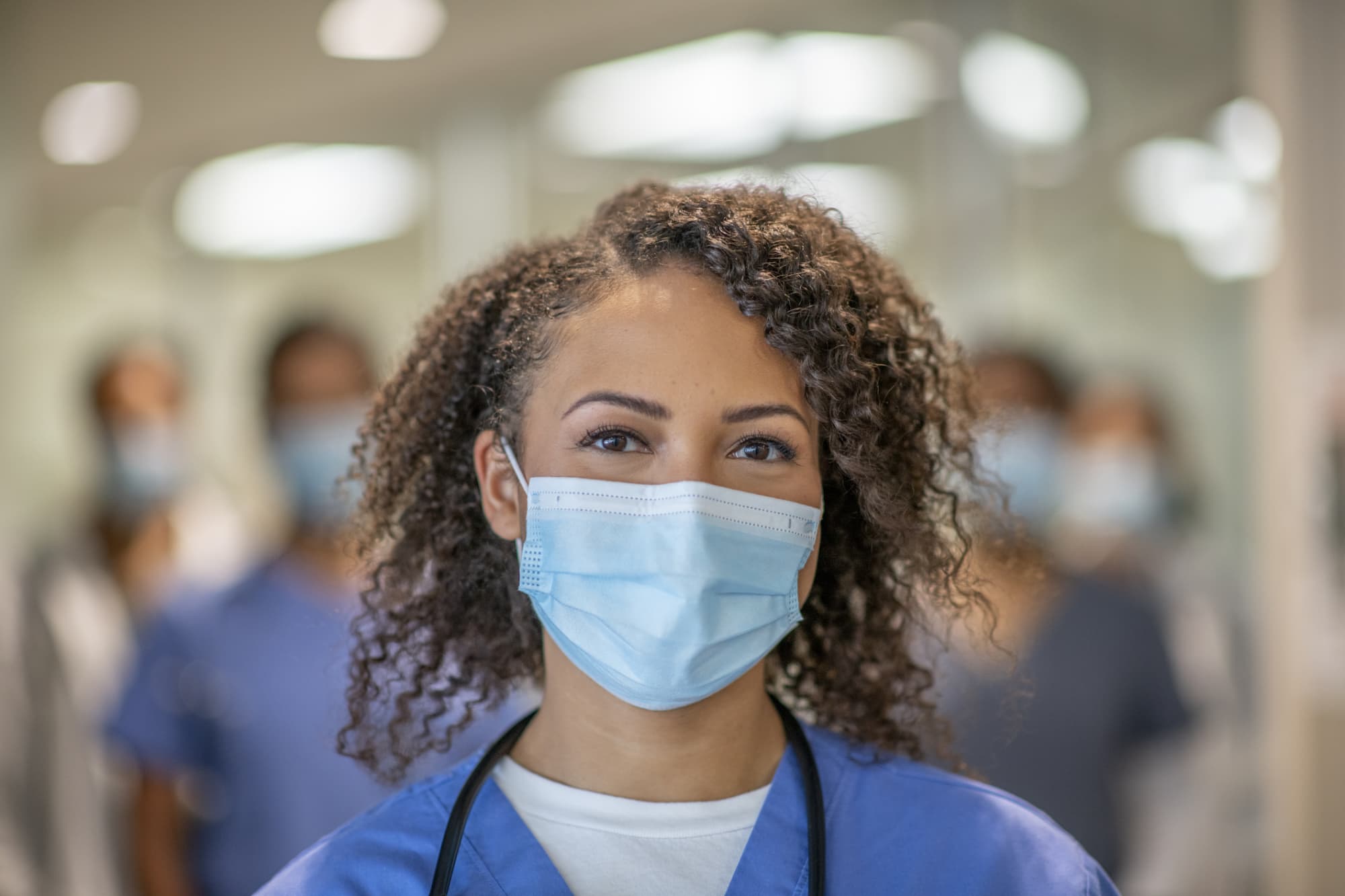 A female medical professional is wearing blue medical scrubs and a face mask stands in front of her colleagues at the hospital. She is smiling behind her mask.