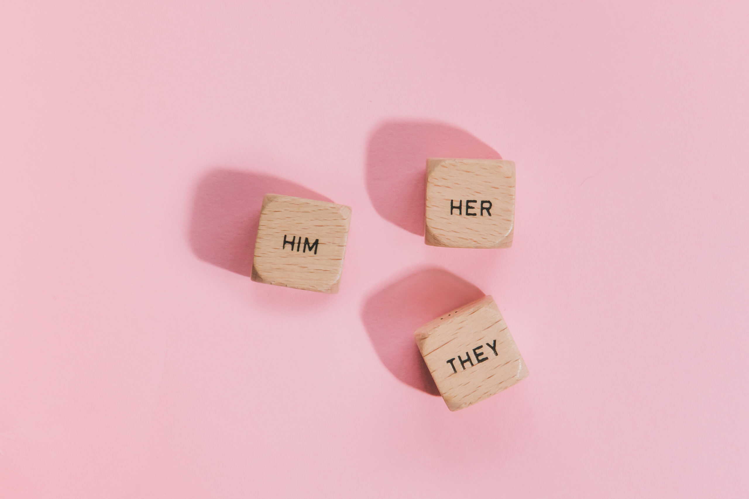 3 wooden blocks laying against a pink background with the words, "him", "her", "they" written on them.