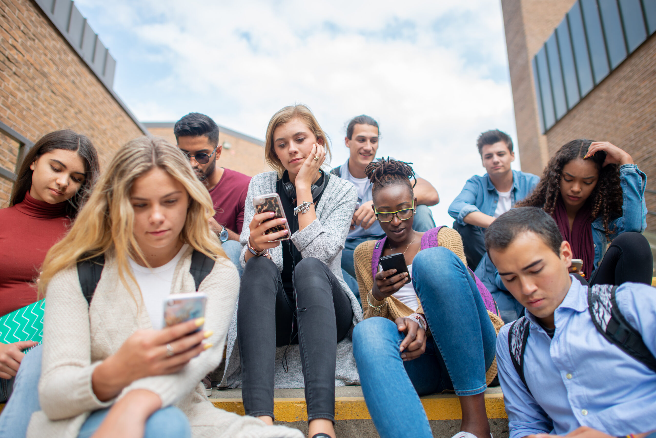 A multi ethnic group of high school students sitting on a flight of stairs outside a school building. They are all busy scrolling on social media on their phones.