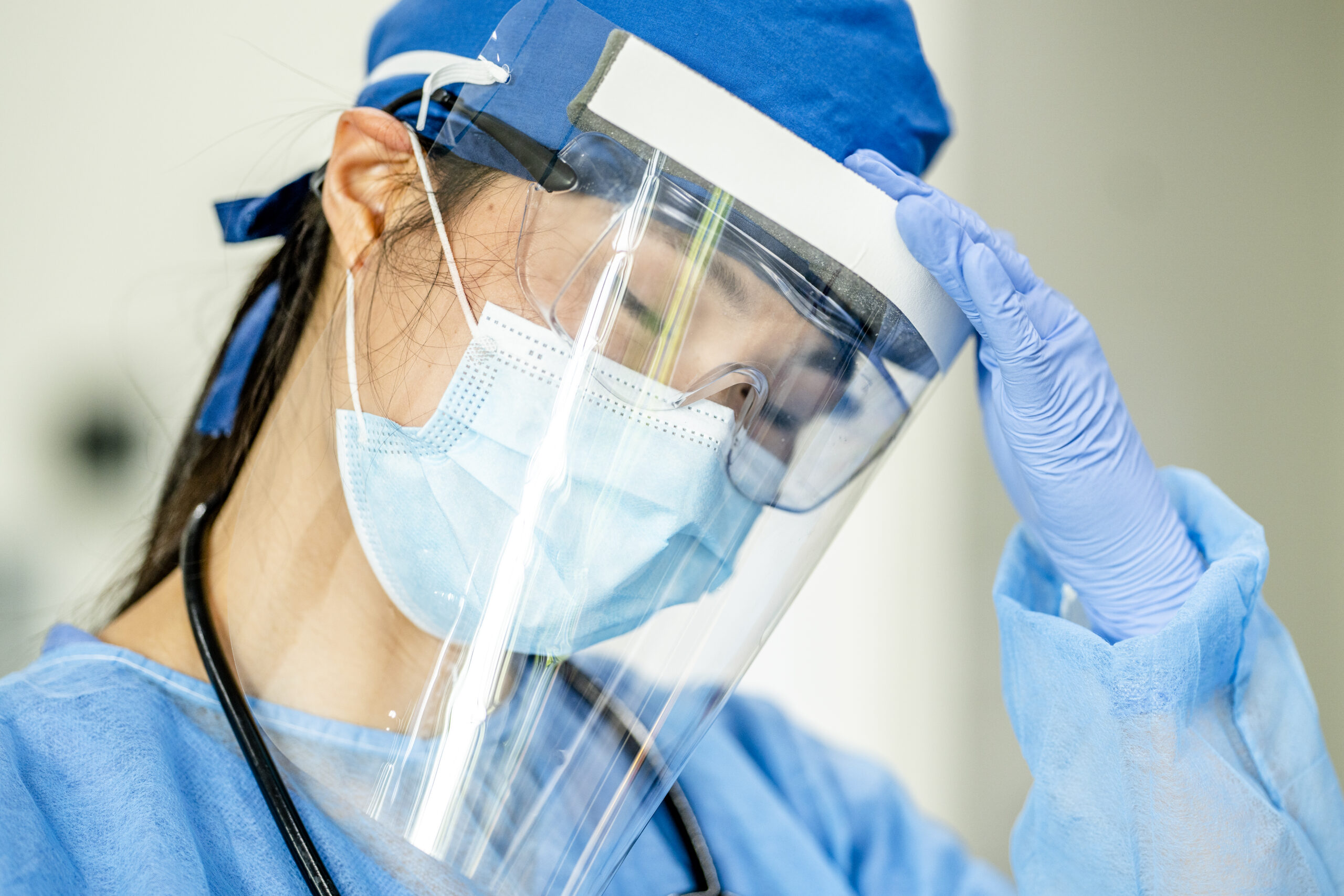 A beautiful female doctor of Asian ethnicity appears to be stressed. She has her hand onto her forehead as though she is having a headache. She is wearing safety goggles, face mask a surgical cap and a face shield.