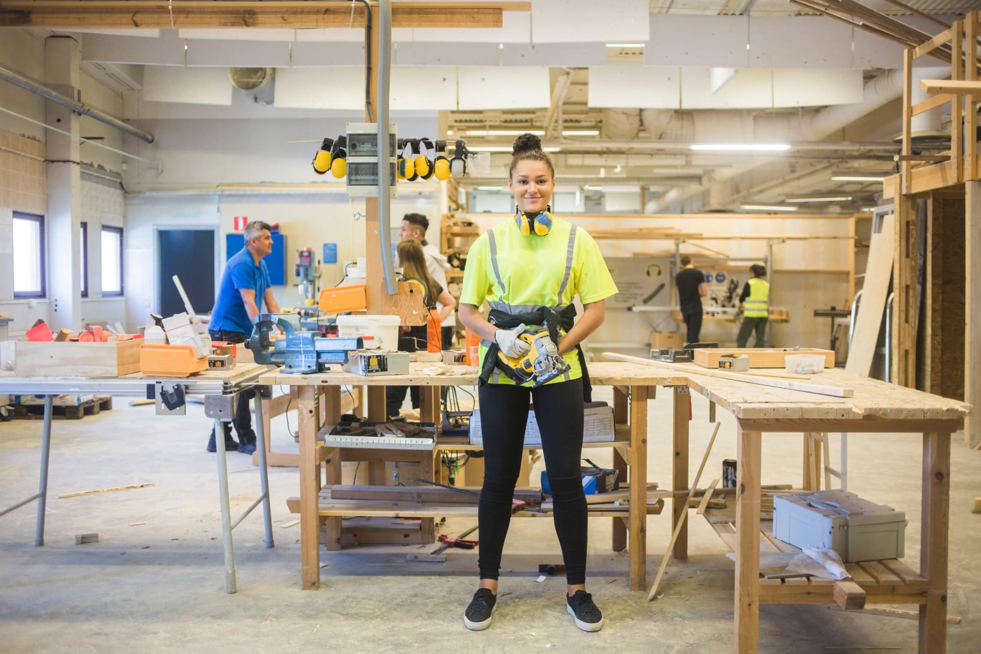 Full length portrait of smiling young female trainee holding power tool while standing by workbench at illuminated works