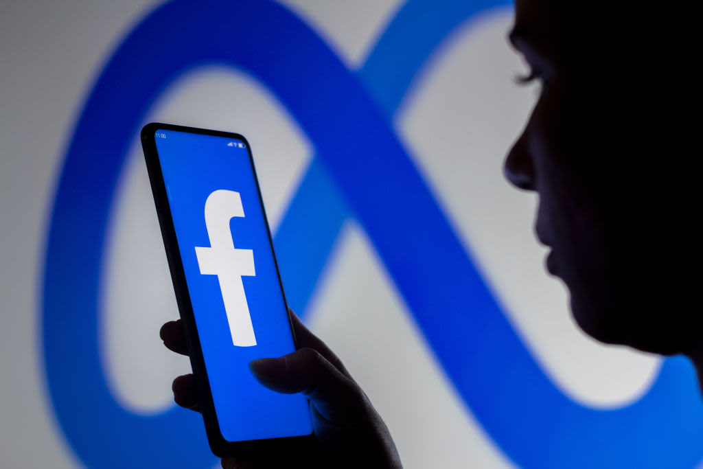 A woman standing in front of a Meta Platforms brand sign holds a smartphone with the Facebook logo displayed on the screen. The Department of Education has been tight-lipped since a report revealed that FAFSA applicant info may have been shared with Meta.