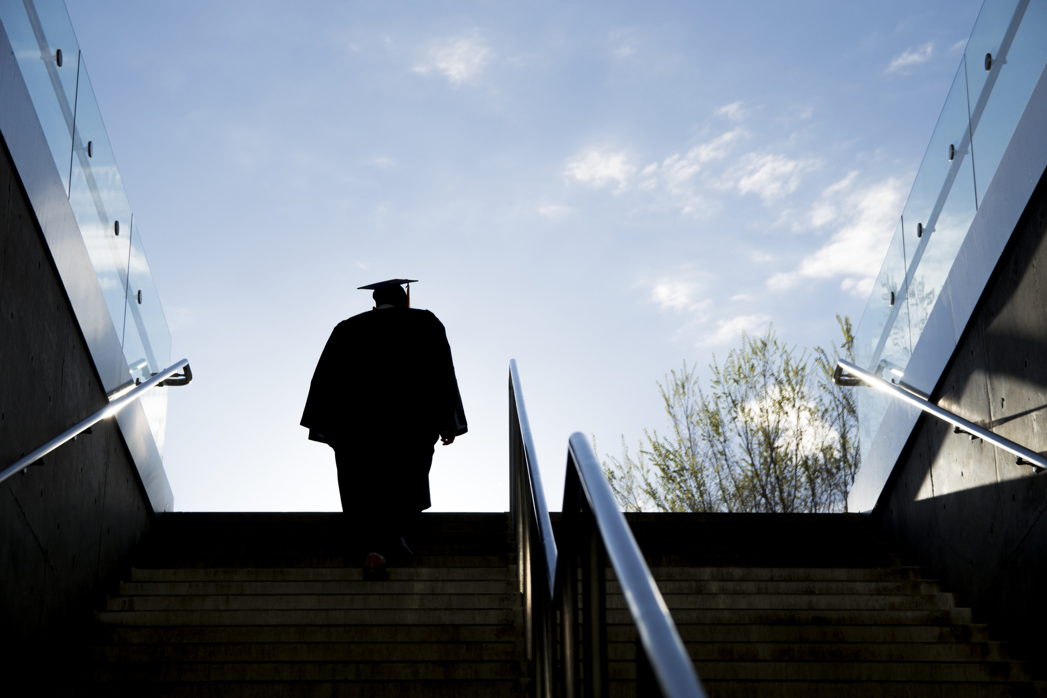 Silhouette of a college graduate in a cap and gown climbing up a set of steps on a university campus.