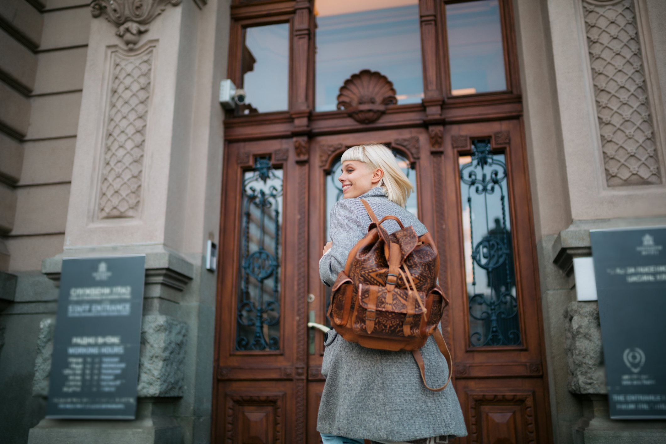 Female university student with blond hair walks towards a building entrance on campus with her backpack and smiles.