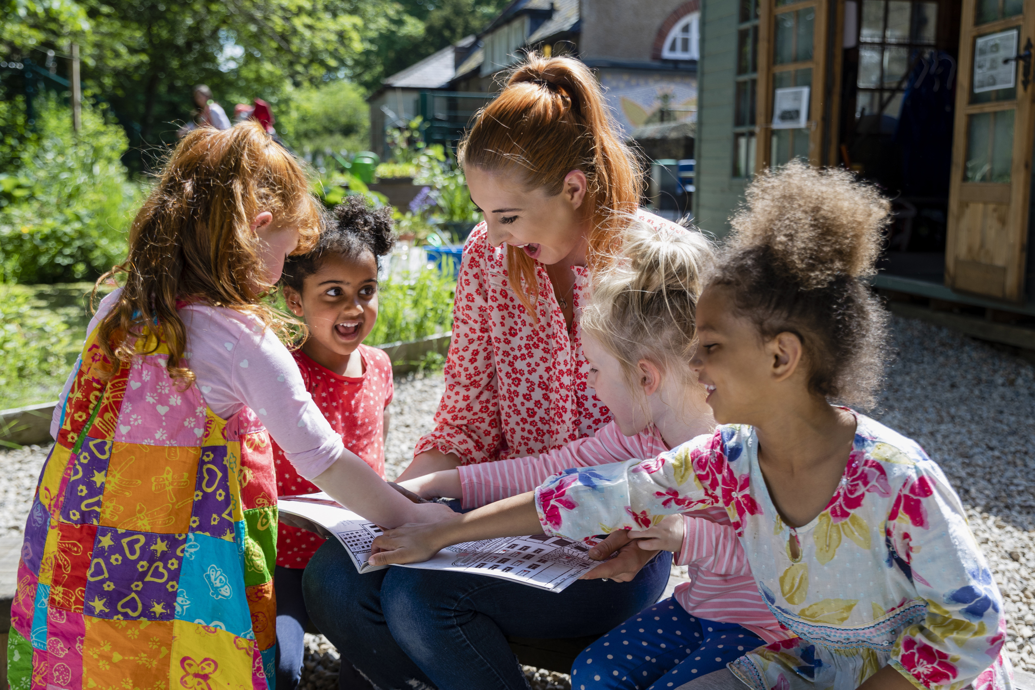 A front view of a teacher and her excited little pupils sitting and doing storytime outdoors in the schoolyard on the benches on a wonderful summer's day.