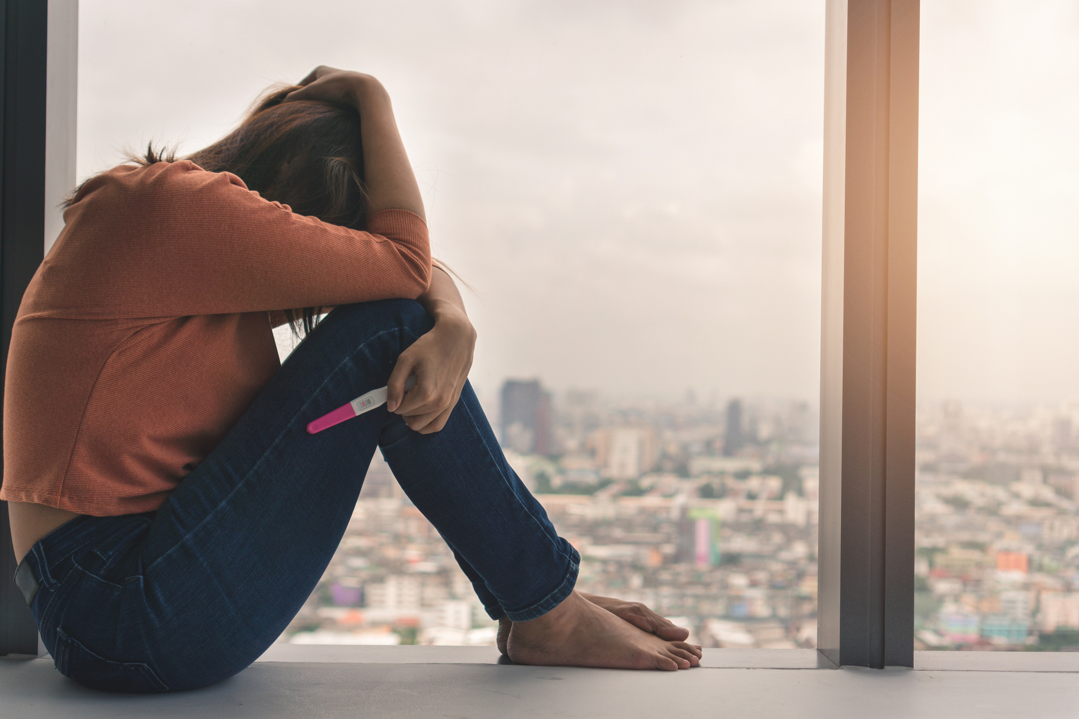 Unhappy young asian woman holding pregnancy test showing a negative result and sit down near big window city view on evening time, Wellness and healthy concept, Infertility problem, Selective focus.