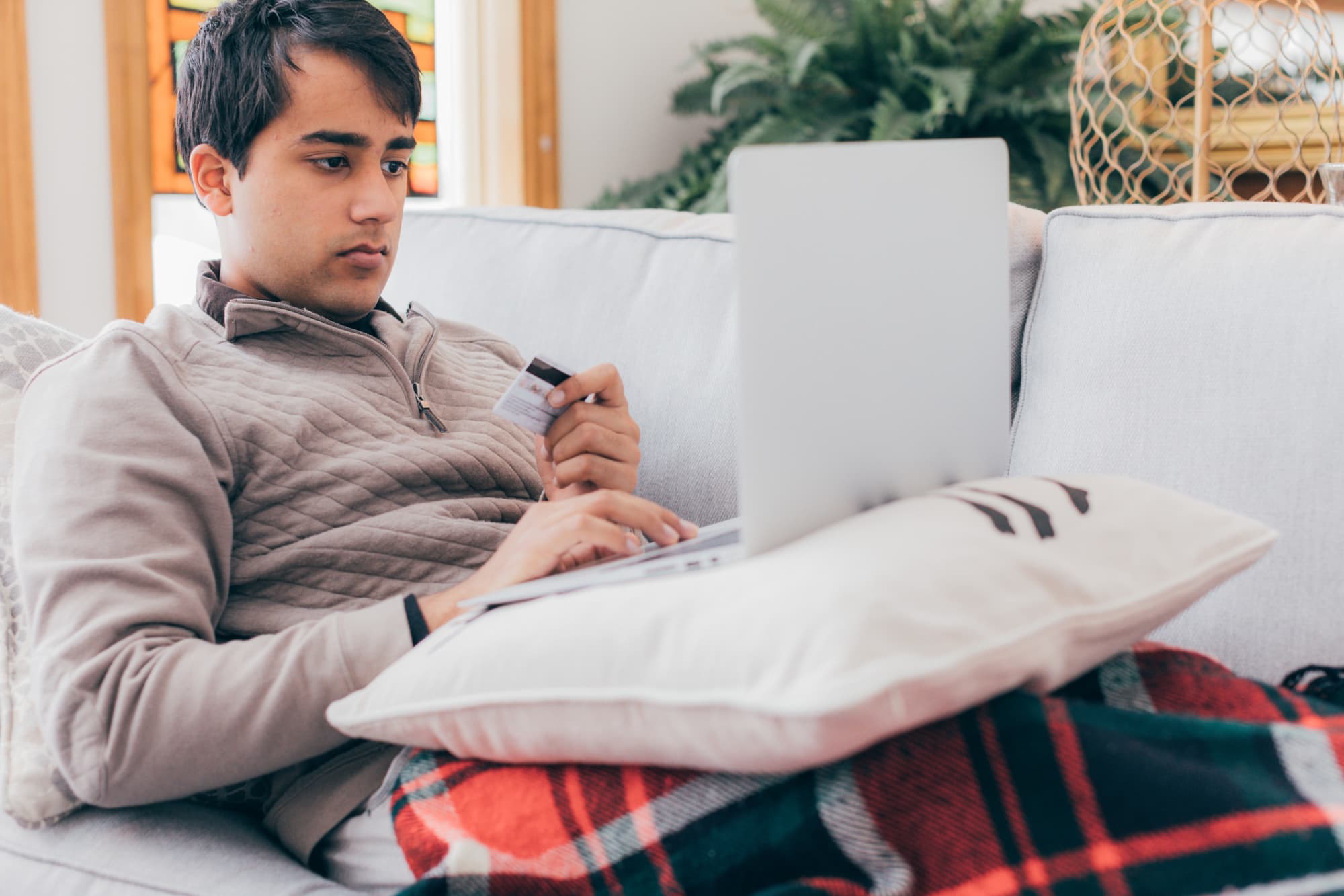 College student lying on a couch in his living room paying his student loan bill online with his credit card.