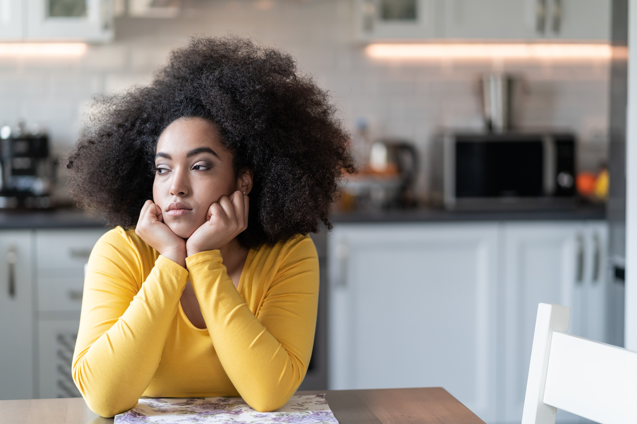 A depressed woman sitting at home alone in her kitchen looing sadly out of a window