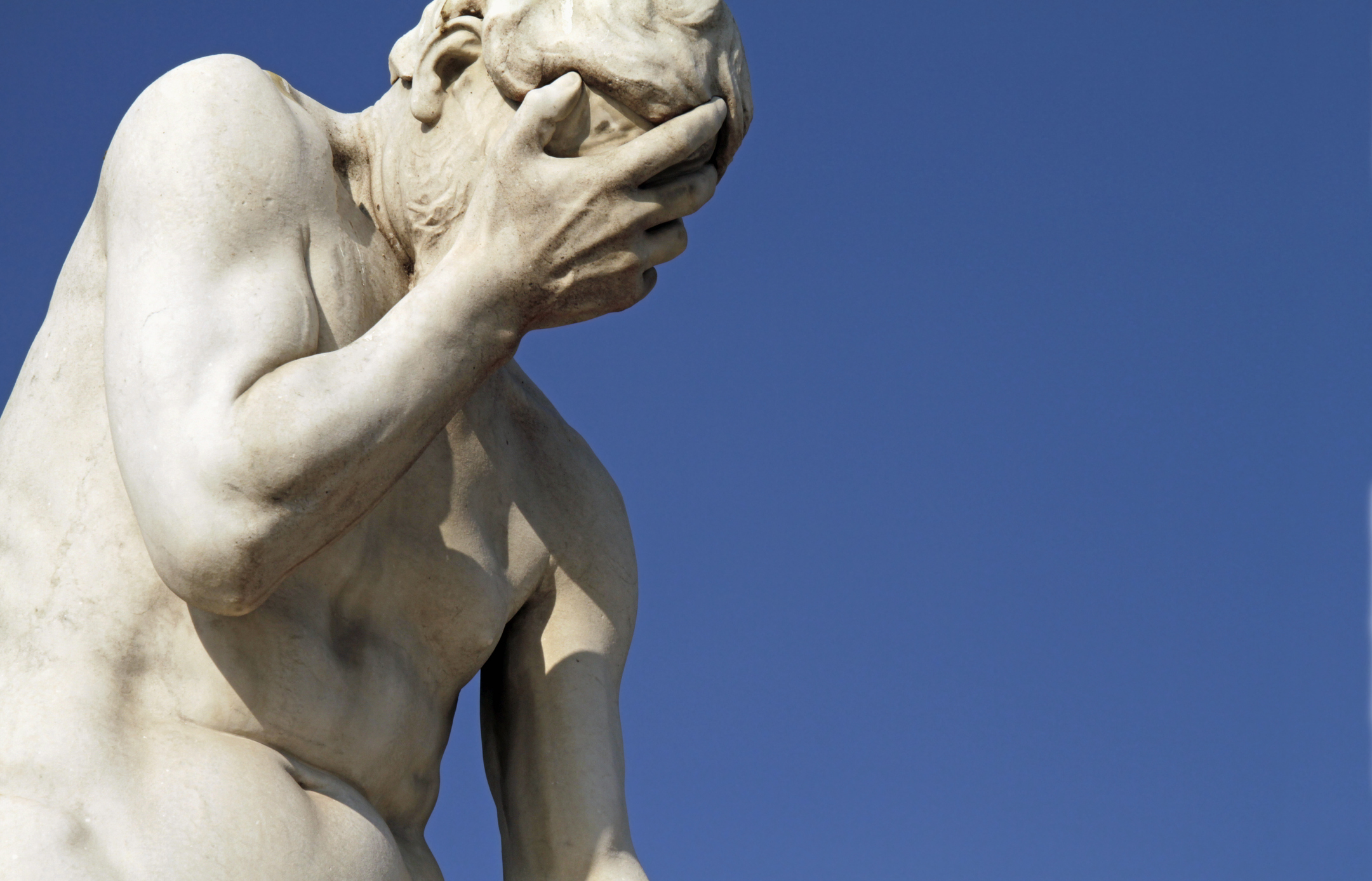 Photo of a marble statue showing a person Facepalming, Head In Hands against a clear blue sky background