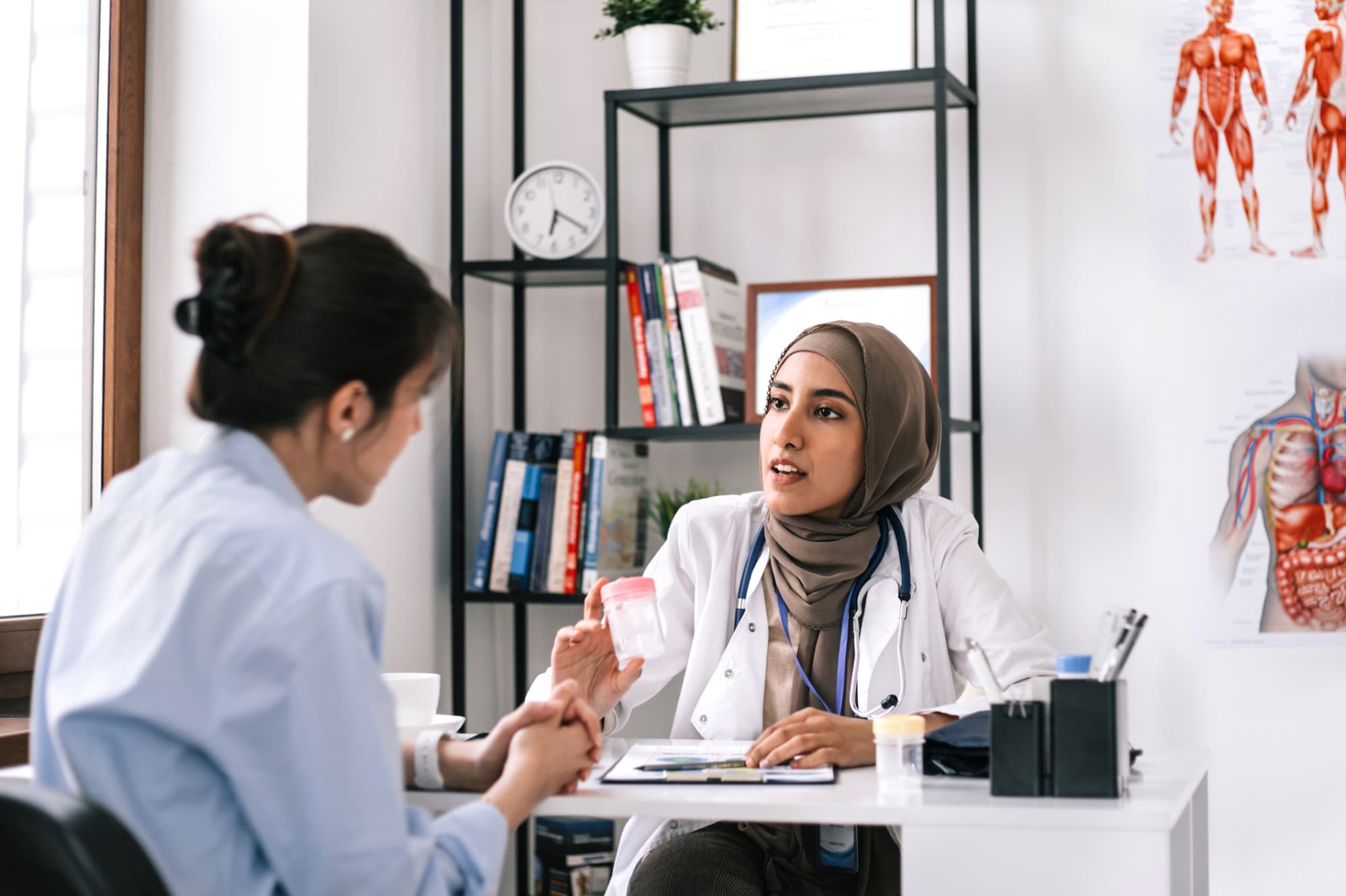 A female Muslim doctor sitting at the desk in her office chats with a female patient during an appointment. The doctor is holding a bottle of prescription medication in her hand and handing it to the patient.