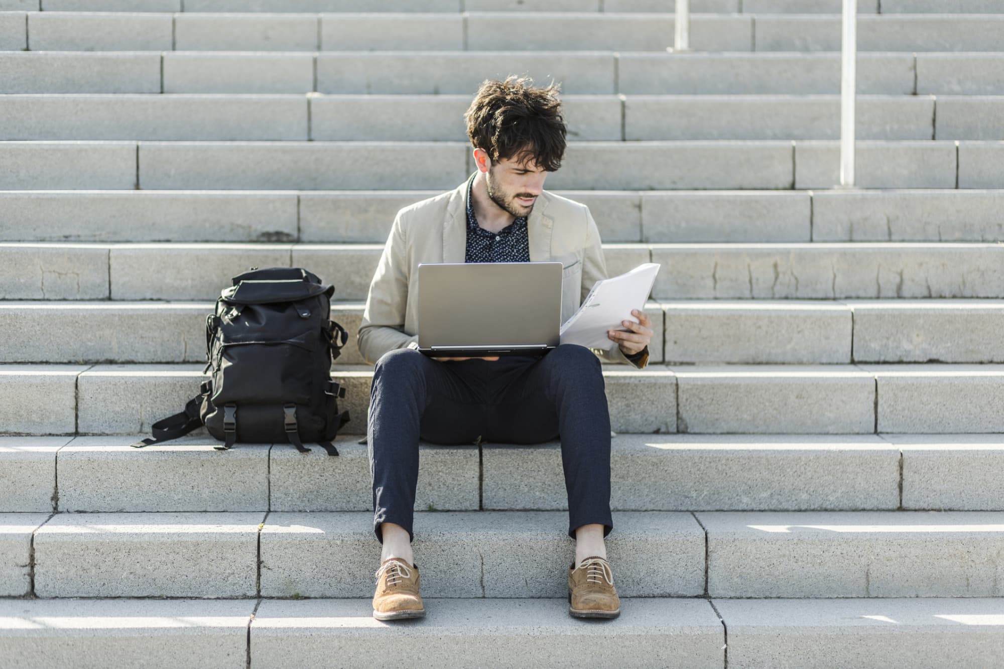 Law student with a back pack and laptop sitting on steps outside a campus building reviews a paper copy of his LSAT score.