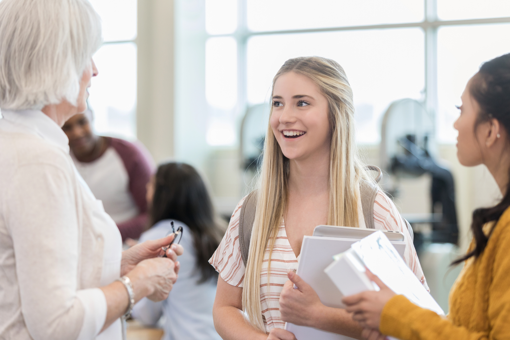 Teenage girl standing in a high school hallway with her friend holding her textbooks in hand. She is talking with a school administrator, who's just informed her that she won a competitive scholarship.