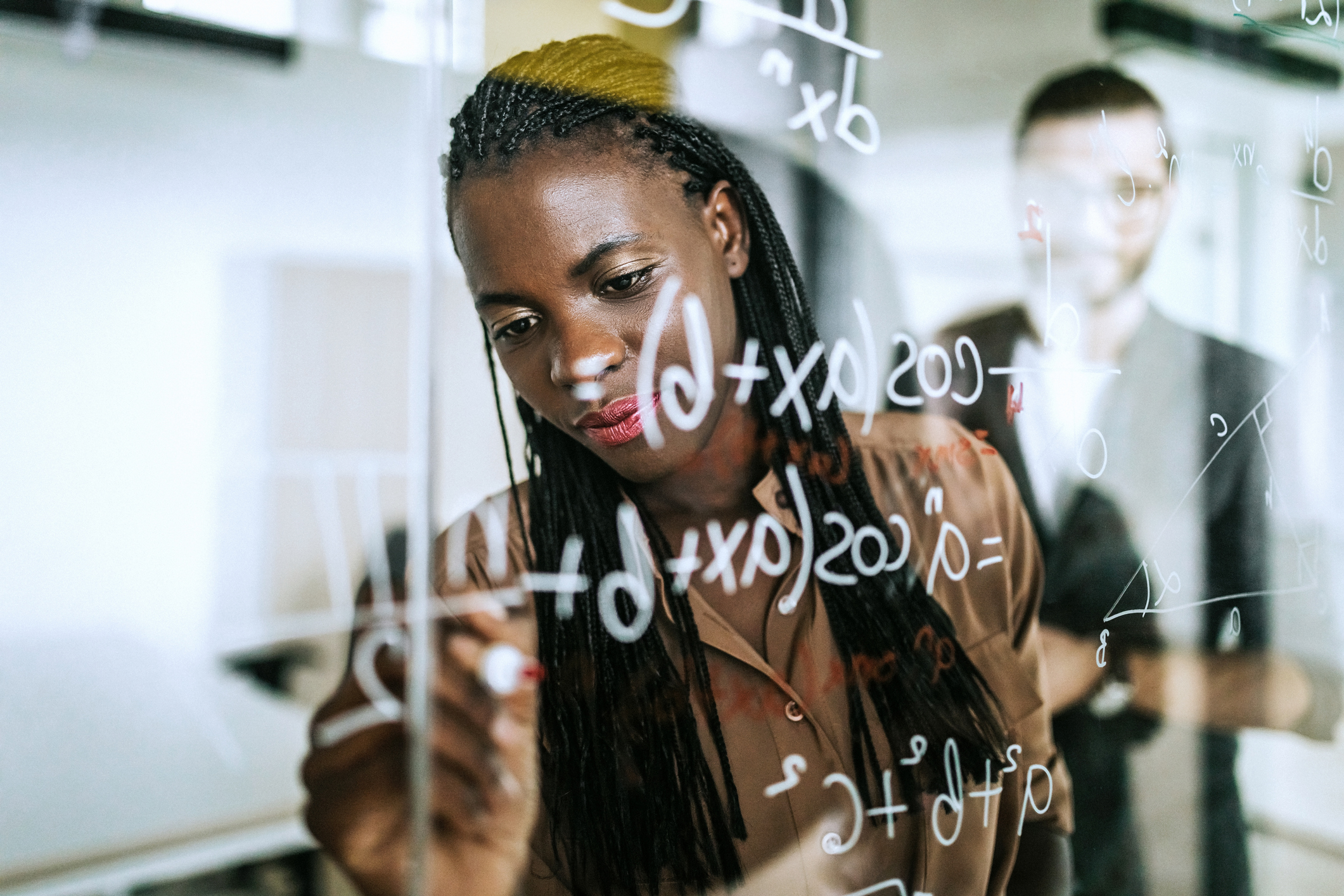 Black female math student writes mathematic formulas with a marker on a transparent wipe board during class.