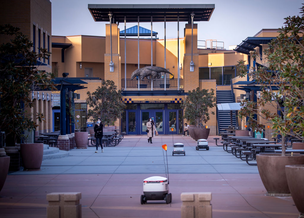 Starship food delivery robots making meal deliveries on the University of California Irvine campus as students walk amidst a mostly empty food court and student center outside.