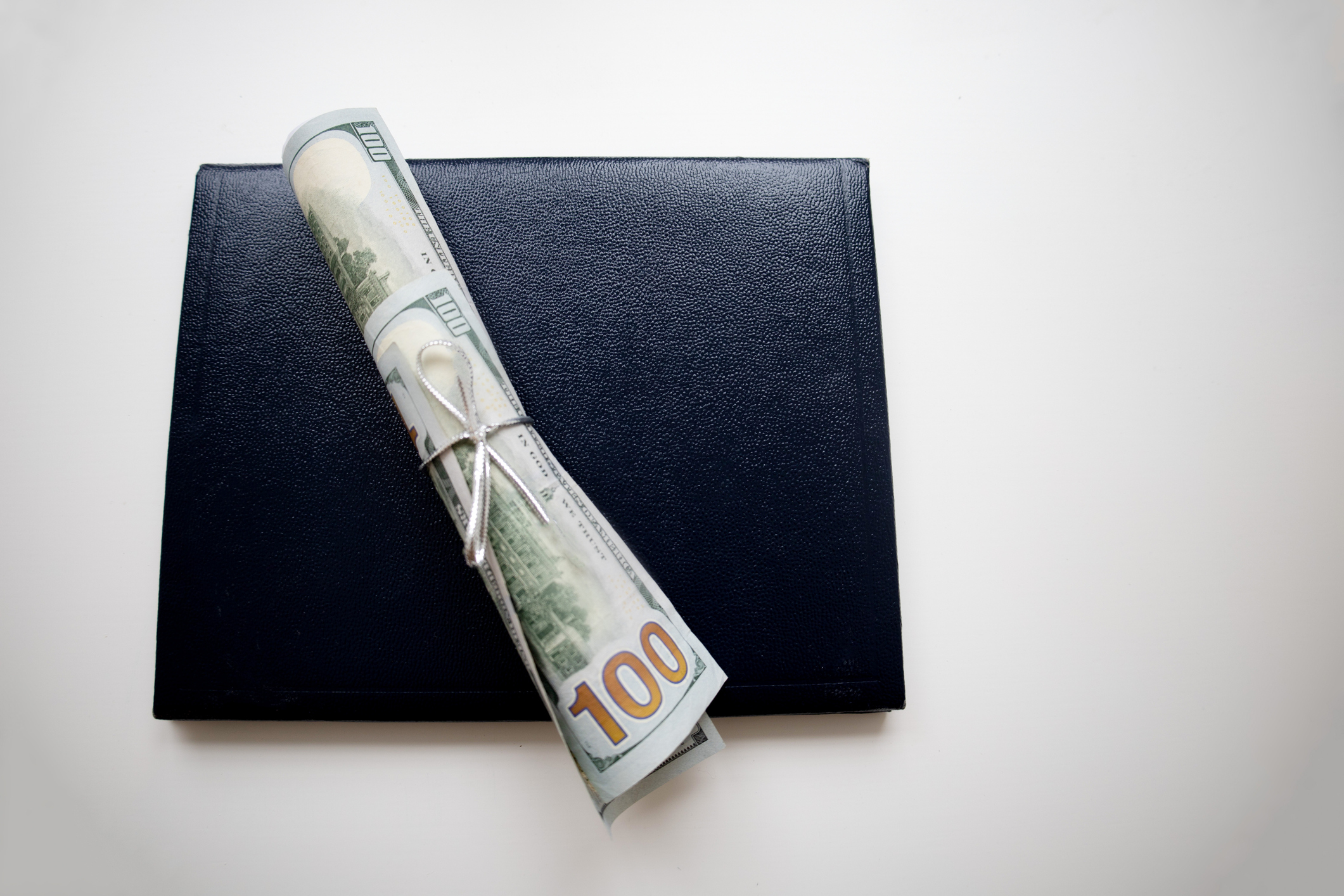 graduation & student loan debt concept image. a one hundred dollar bill folded up like a diploma, rests on top of a graduation cap. Top down photo against a gay background.