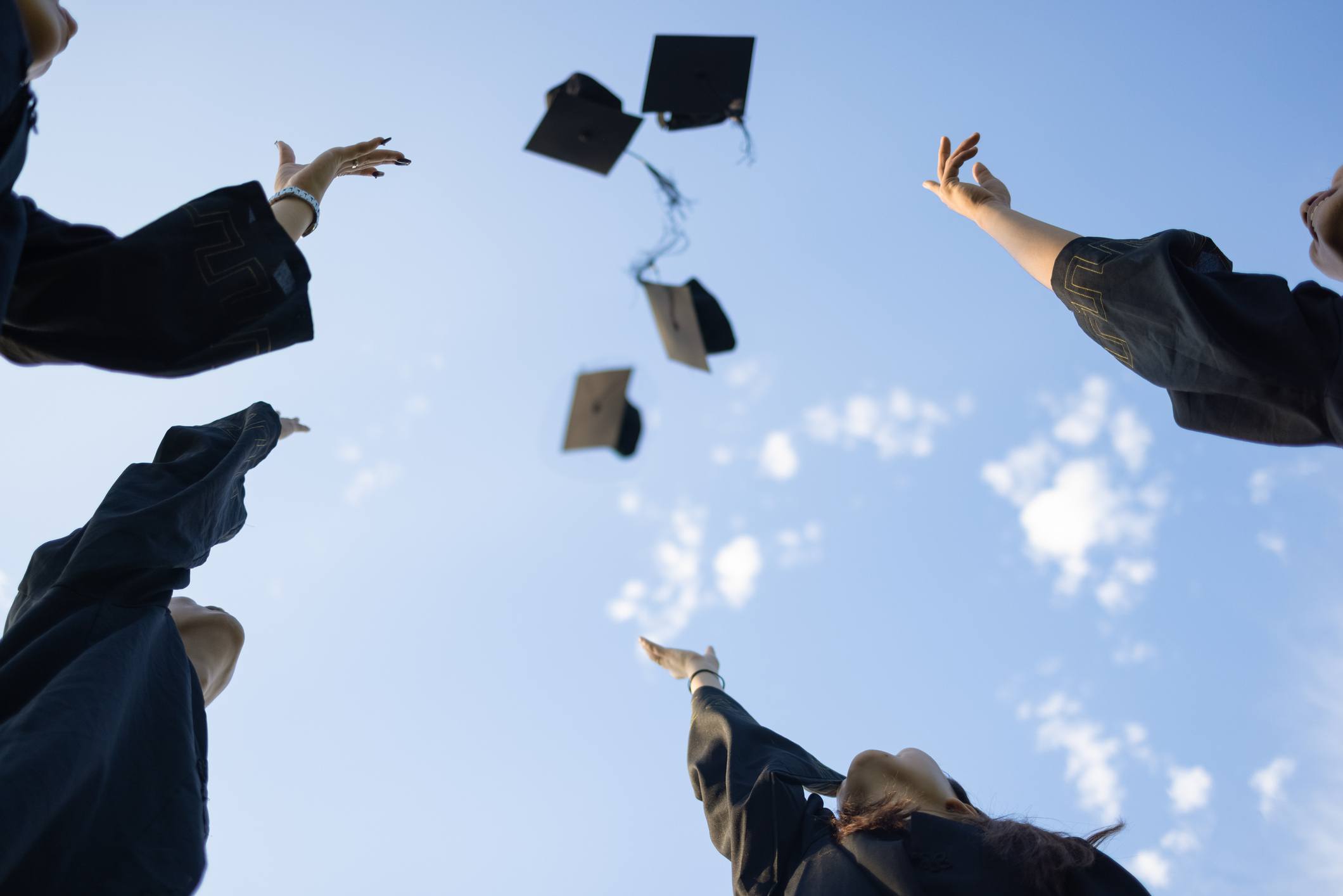 A group of college students dressed in black graduation gowns celebrate outside after their graduation ceremony by throwing their caps into the air.