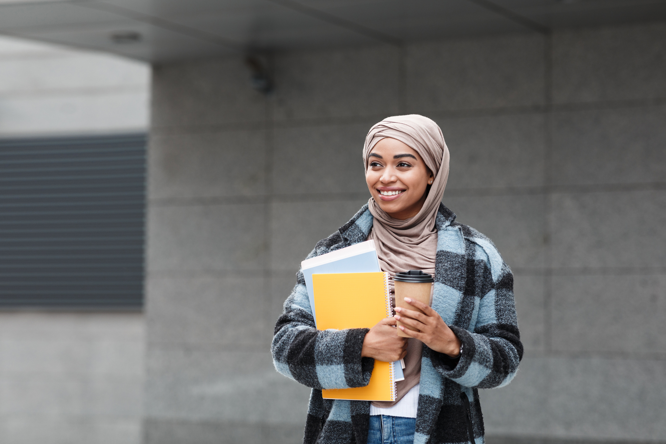 A first-generation Black female student wearing a hijab scarf and casual Western coat and jeans walks to class on campus. She is holding a pair of notebooks in her right arm and a cup of coffee in her left. She is smiling and looking happy.