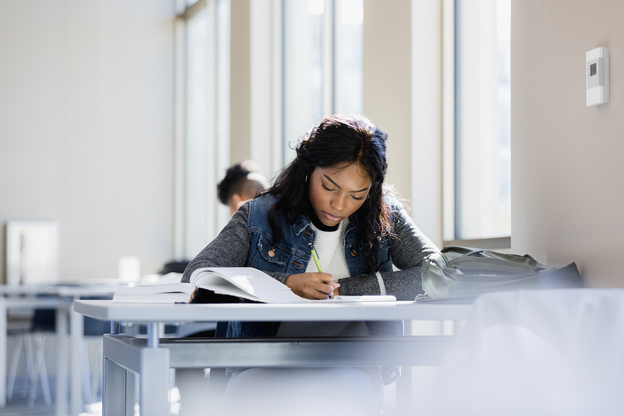 Black female college student sitting at a table in a campus library. She focused on taking notes while reading through her textbook.