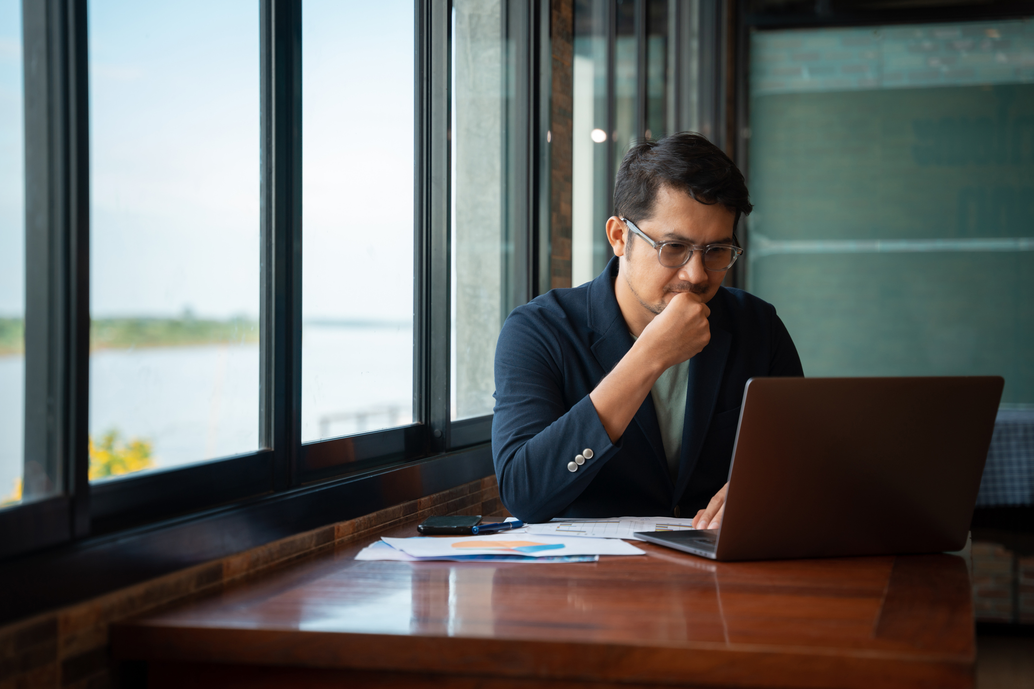 Asian American male Ph.D. student sitting at a table in a cafe. He is typing a report on his laptop computer. There are several paper notes on the table that he is referencing.